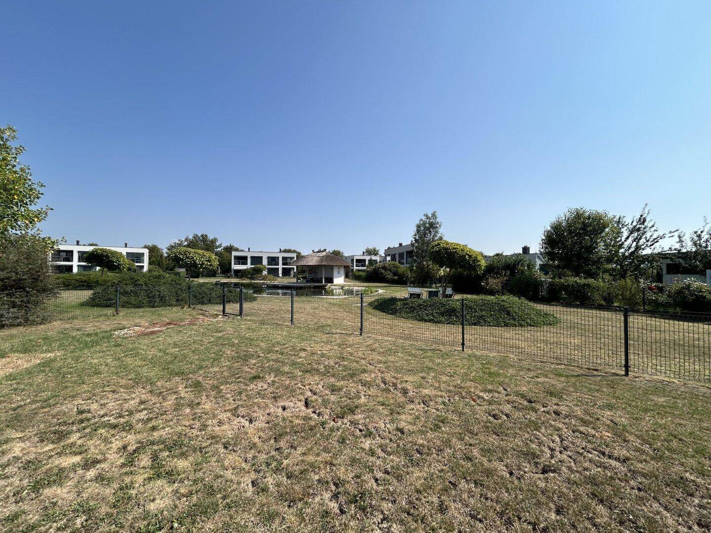 Weitläufiger Gemeinschaftsgarten mit Rasenflächen, Bäumen und einem kleinen Pavillon am Teich.