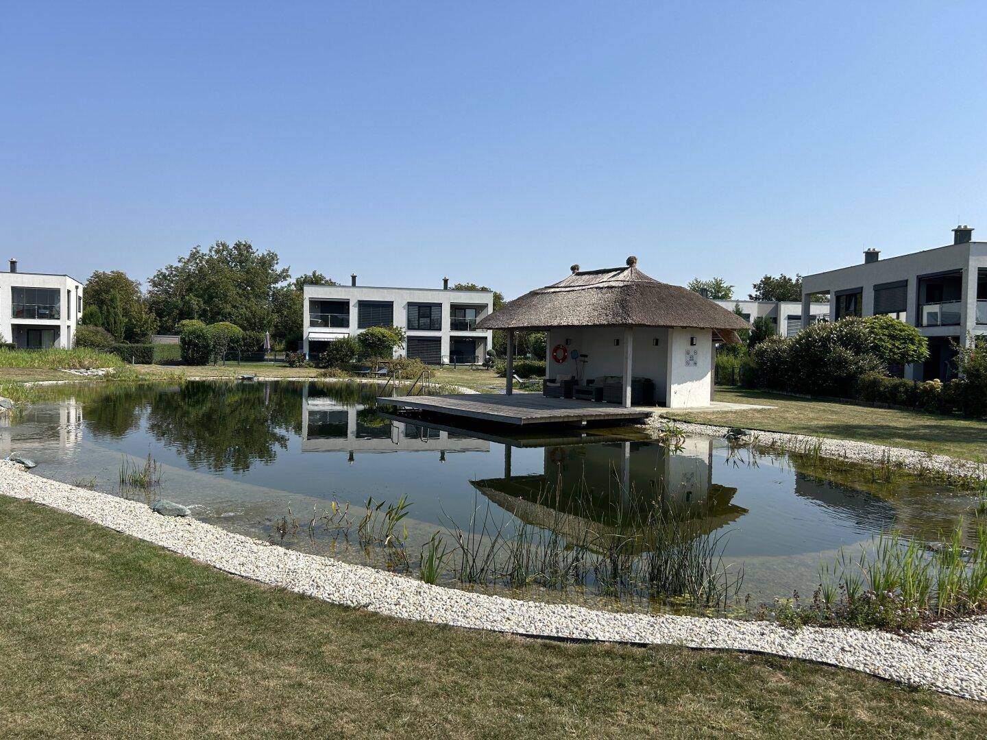 Idyllischer Teichbereich im Garten mit einer kleinen Hütte und einer Holzplattform, umgeben von Grün.
