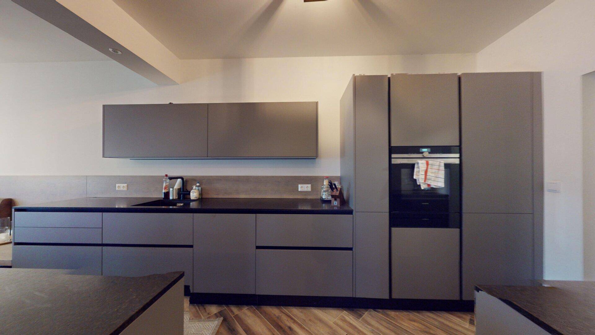 Moderne Einbauküche mit grauen Fronten, dunklen Arbeitsplatten und integriertem Backofen.