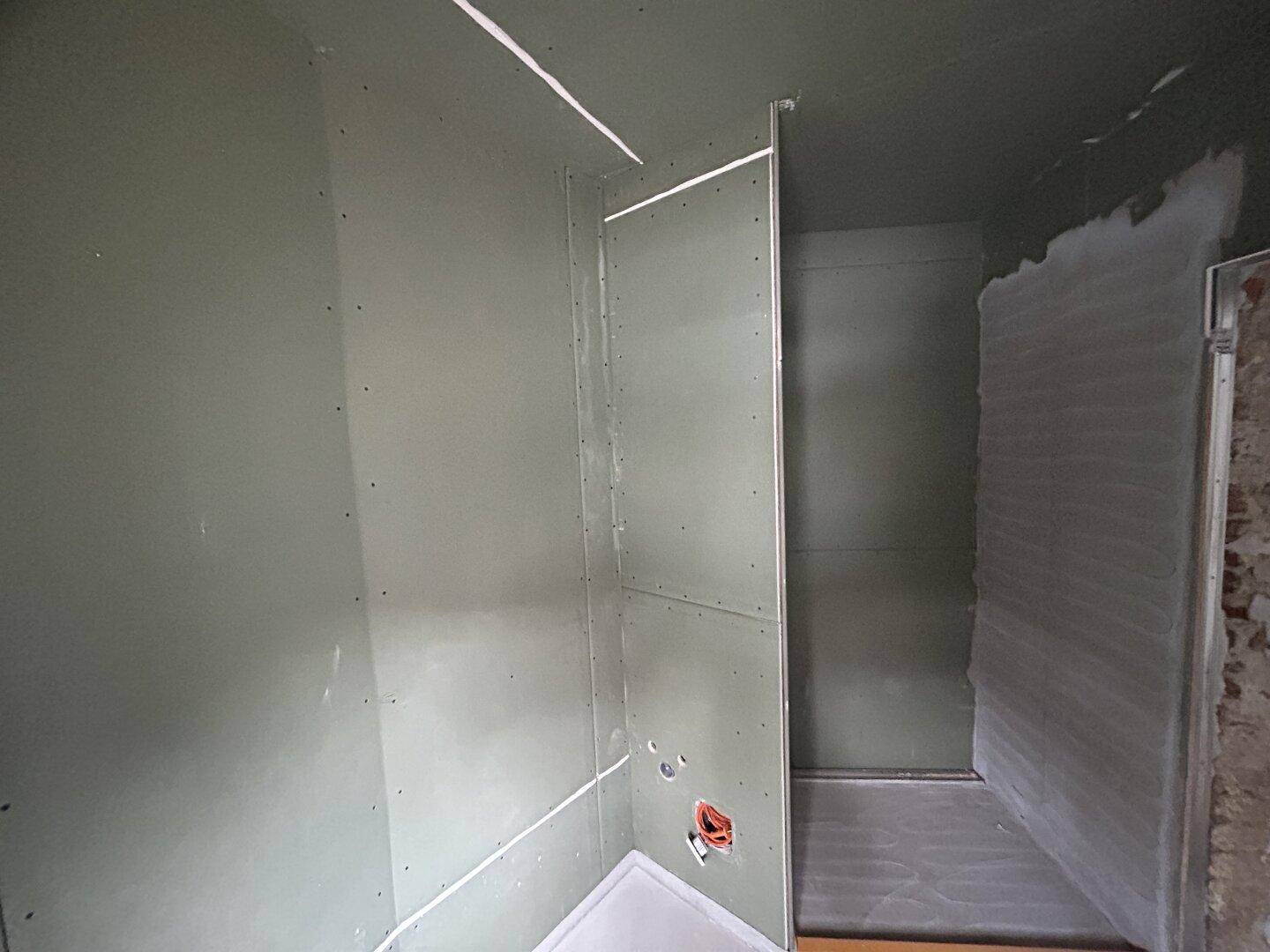 Badezimmer im Rohbau mit vorbereiteter Duschfläche und Wandverkleidung.