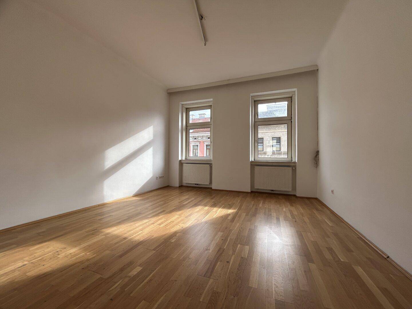 Geräumiger, heller Raum mit zwei Fenstern, Holzfußboden und viel natürlichem Sonnenlicht.