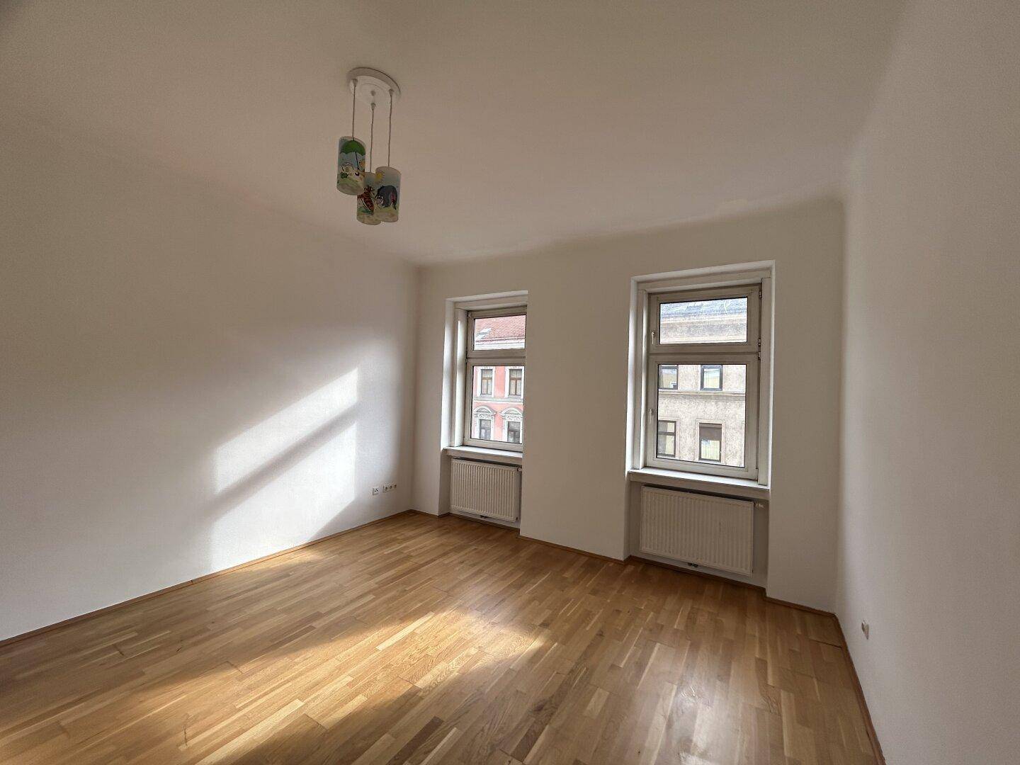 Geräumiger, heller Raum mit zwei Fenstern, Holzfußboden und einer Deckenleuchte.