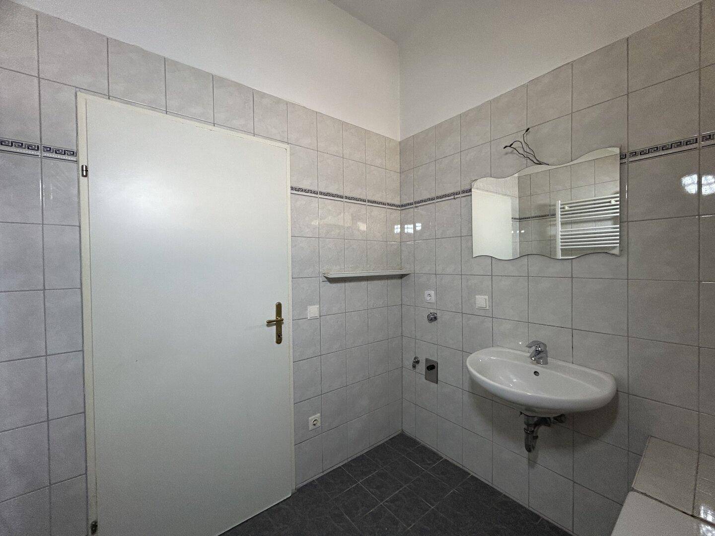 Badezimmer mit Waschbecken, Spiegel und hellgrauen Fliesen an Wänden und Boden.