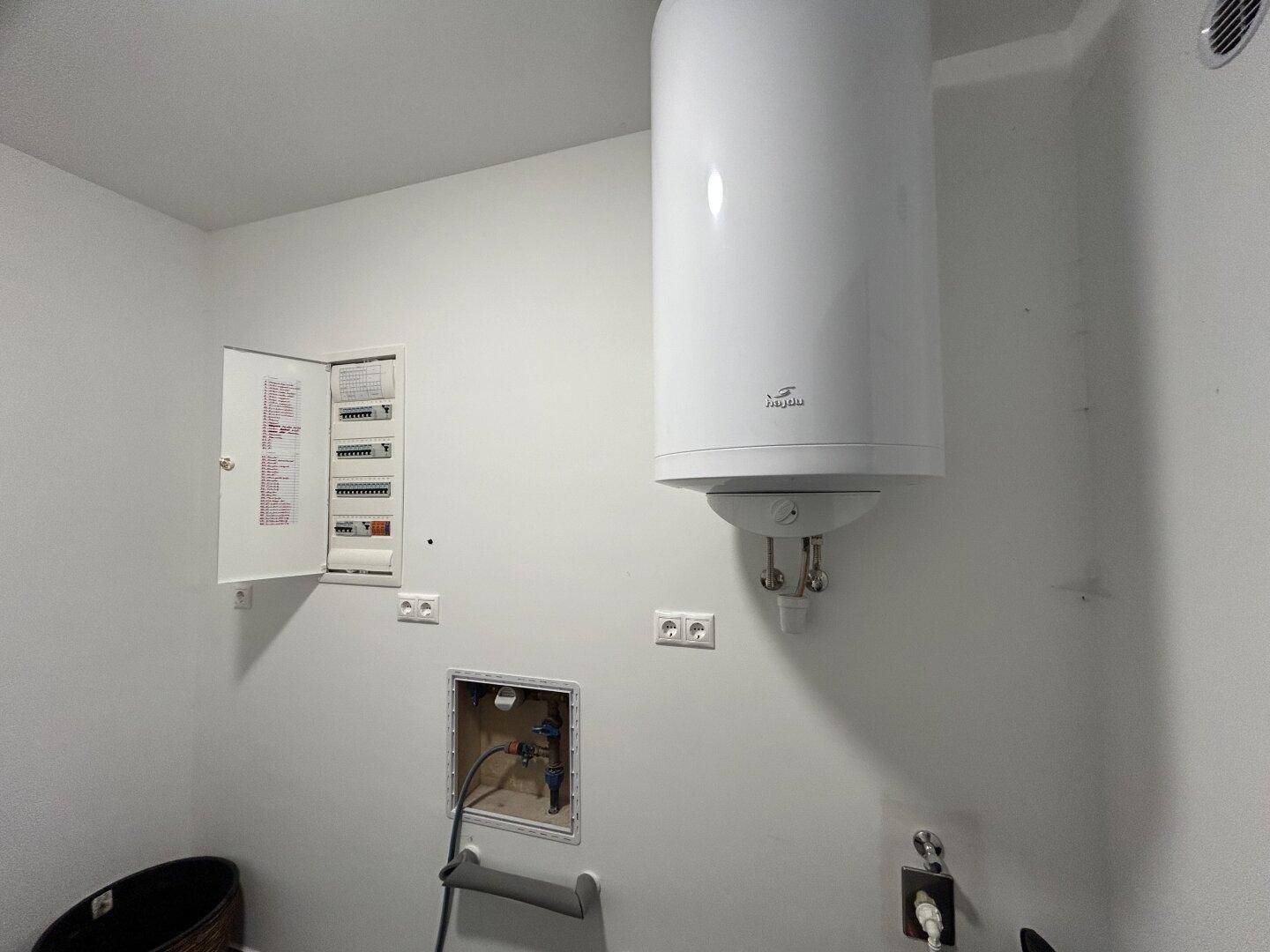 Hauswirtschaftsraum mit Warmwasserboiler, Sicherungskasten und Anschlüssen an weißer Wand.