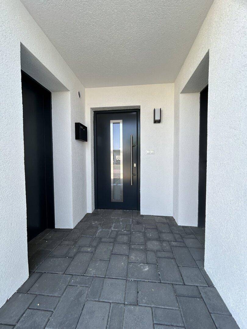 Moderner Eingangsbereich mit dunkler Haustür, schmalem Fenster und grauen Pflastersteinen.