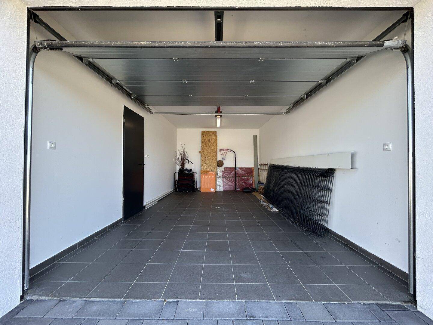 Geräumige Garage mit Fliesenboden, automatischem Tor und Stauraum für verschiedene Gegenstände.