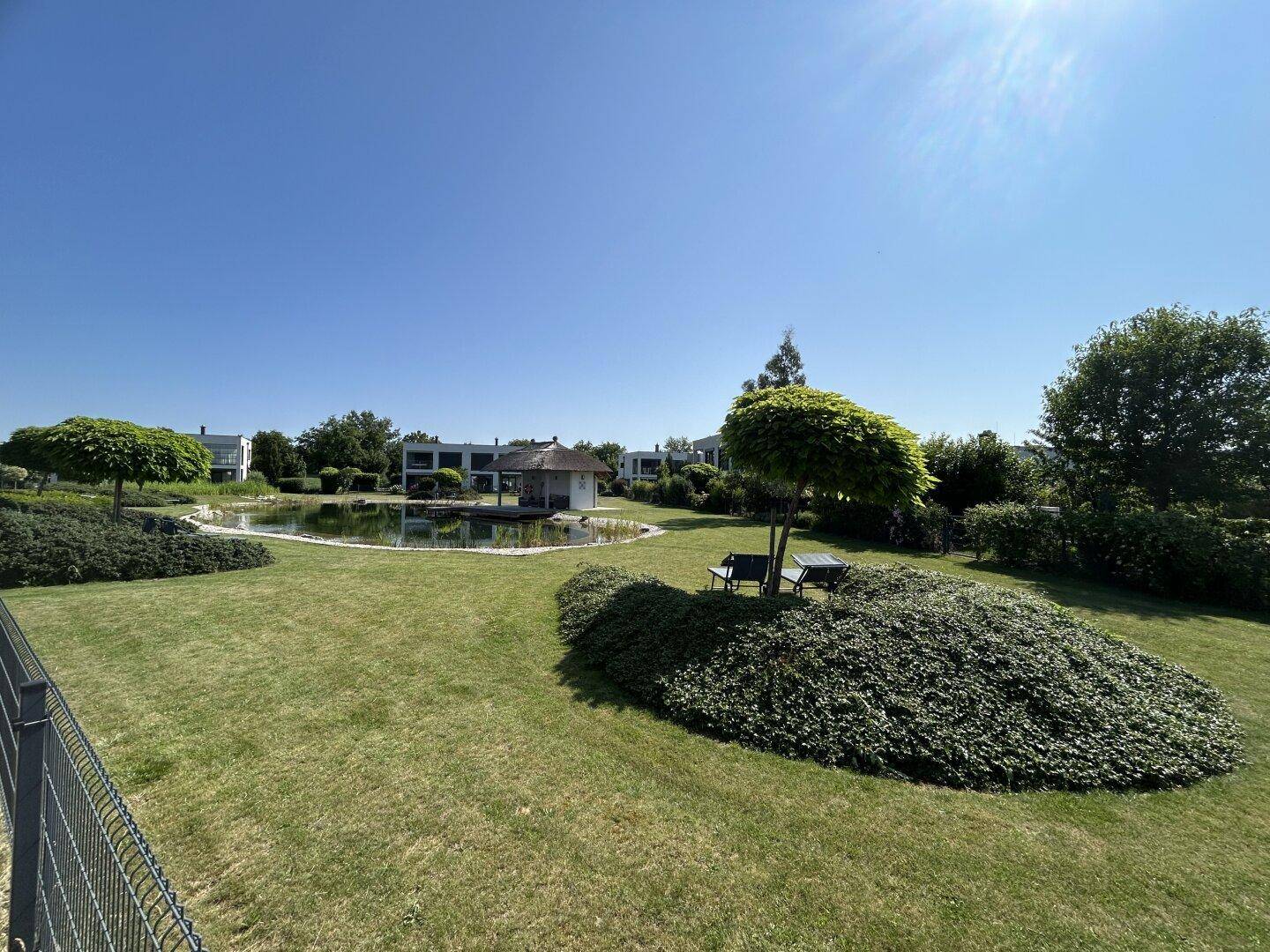 Weitläufiger Landschaftsgarten mit einem natürlichen Teich und modernen Gebäuden im Hintergrund.