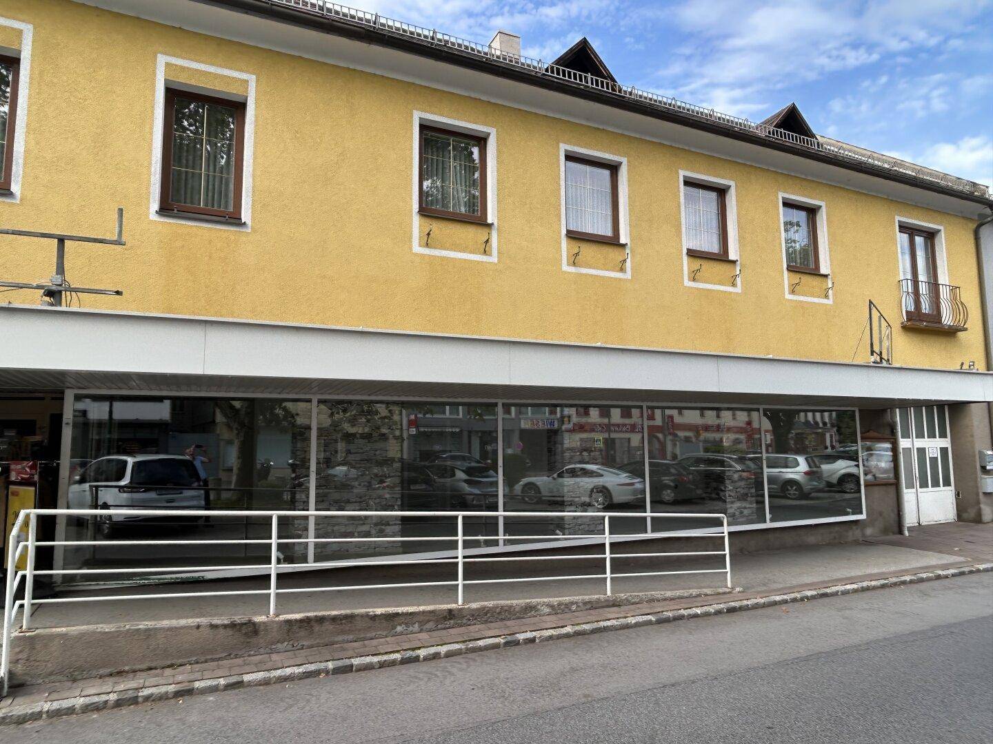 Ansicht der gelben Gebäudefassade mit großen Schaufenstern im Erdgeschoss und einer Zugangsrampe.