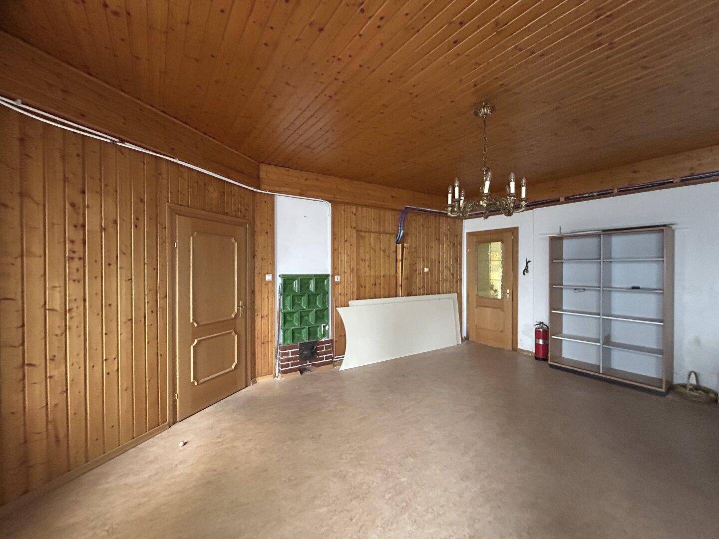 Raum mit Holzvertäfelung, Holzbalkendecke und einem traditionellen Kachelofen, der renoviert werden muss.