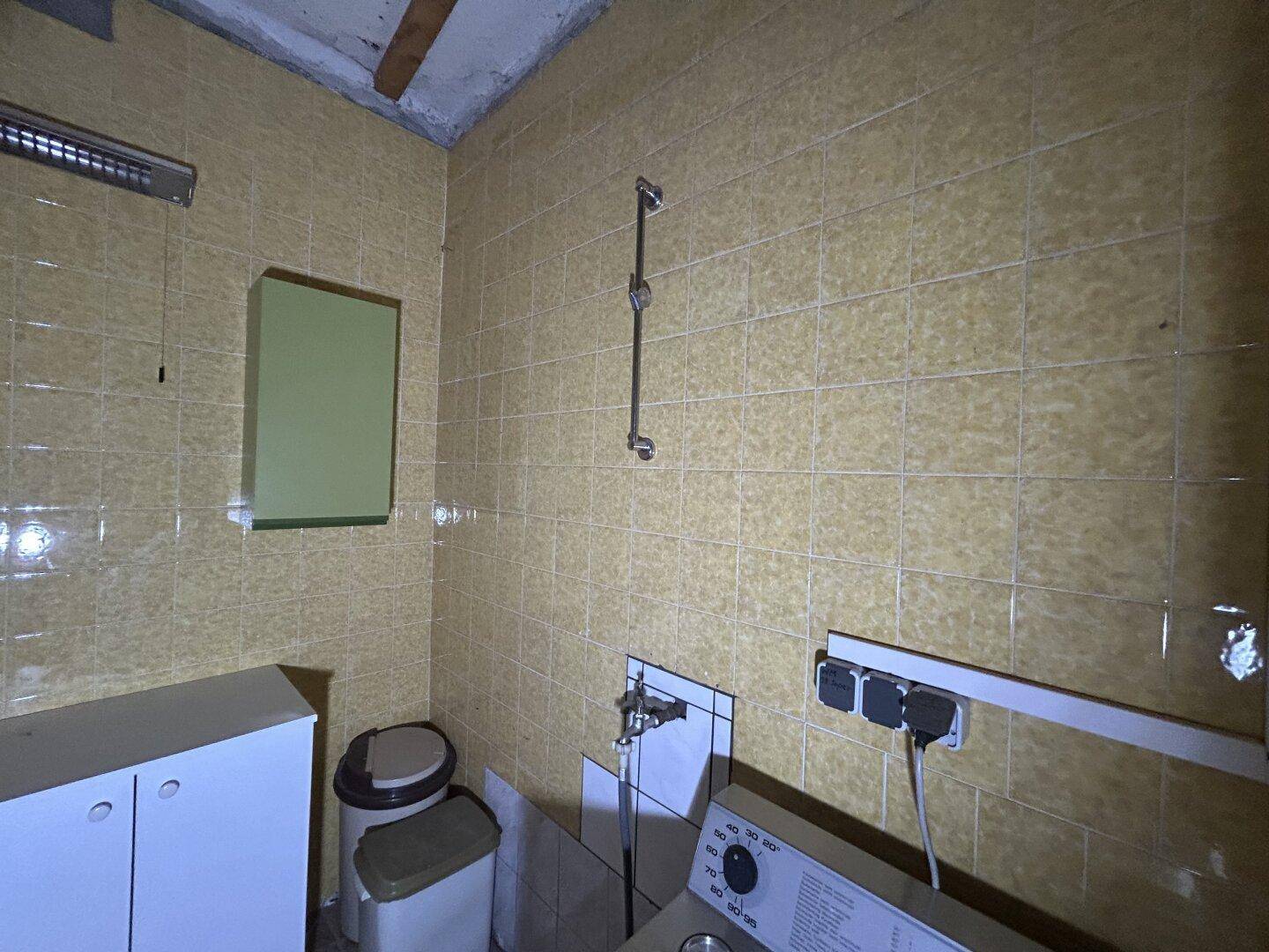 Badezimmer mit gelben Fliesen, Duscharmatur und Waschmaschinenanschluss, das renovierungsbedürftig ist.