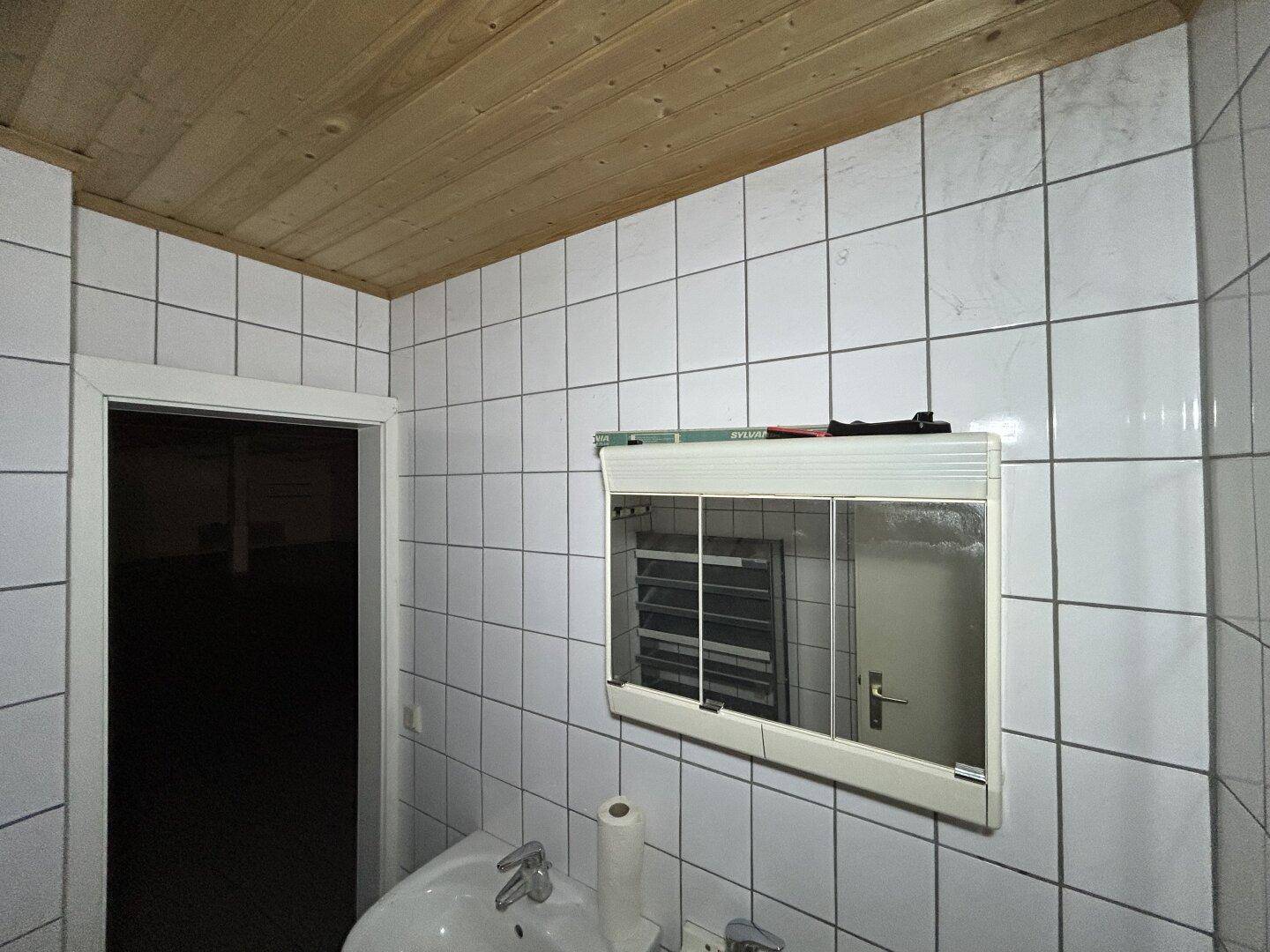 Badezimmer mit weißen Fliesen, Waschbecken und Spiegelschrank, mit Holzdecke.