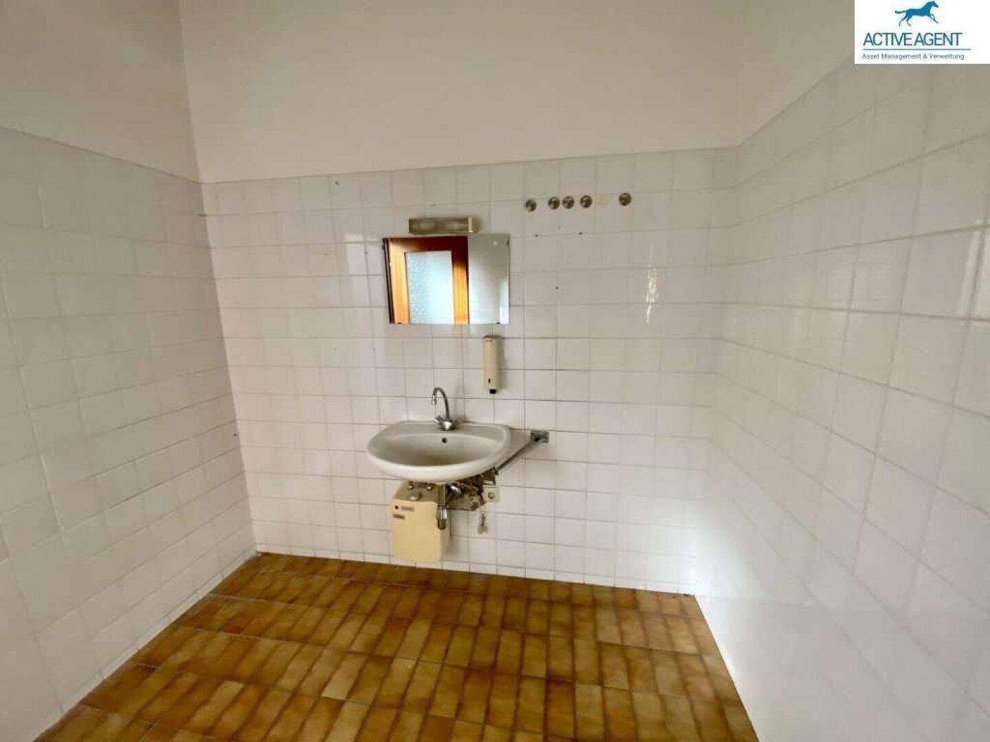 Einfacher Waschraum mit gefliesten Wänden, Waschbecken und einem Spiegel.