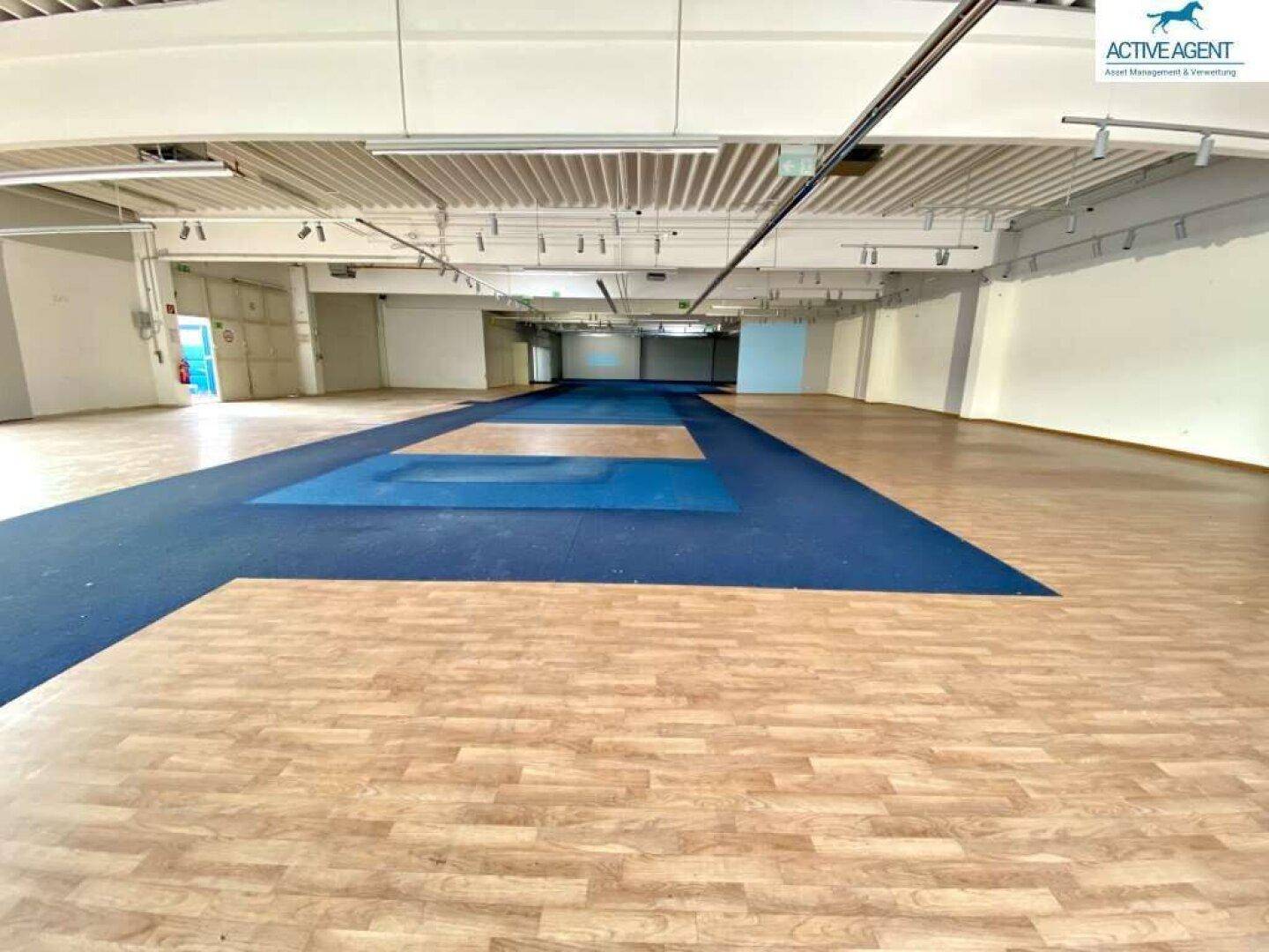 Weitläufiger, leerer Verkaufsraum mit unterschiedlichen Bodenbelägen und hoher Decke.