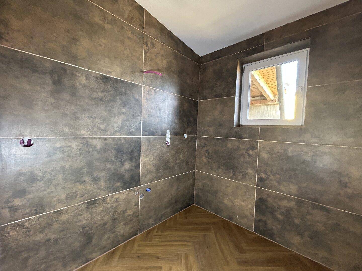 Badezimmer im Bau mit dunklen Fliesen und einem Fenster, das Tageslicht hereinlässt und eine angenehme Atmosphäre schafft.