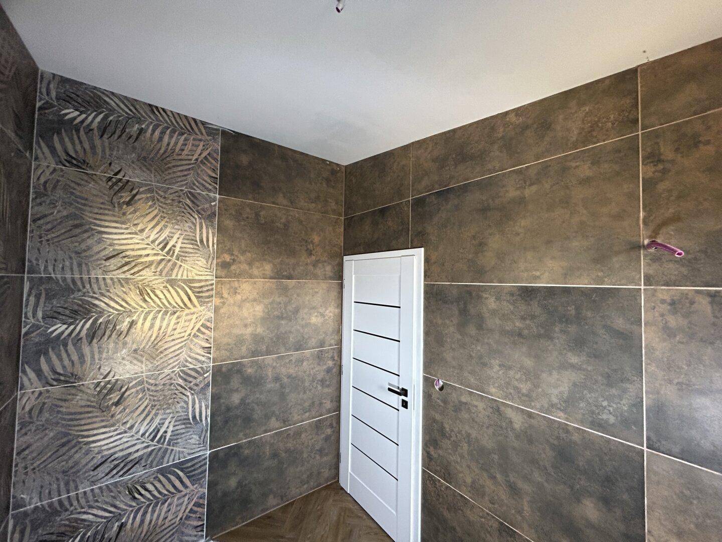 Badezimmer im Bau mit dunklen Fliesen und einer Akzentwand mit Blattmuster, sowie einer modernen Tür.