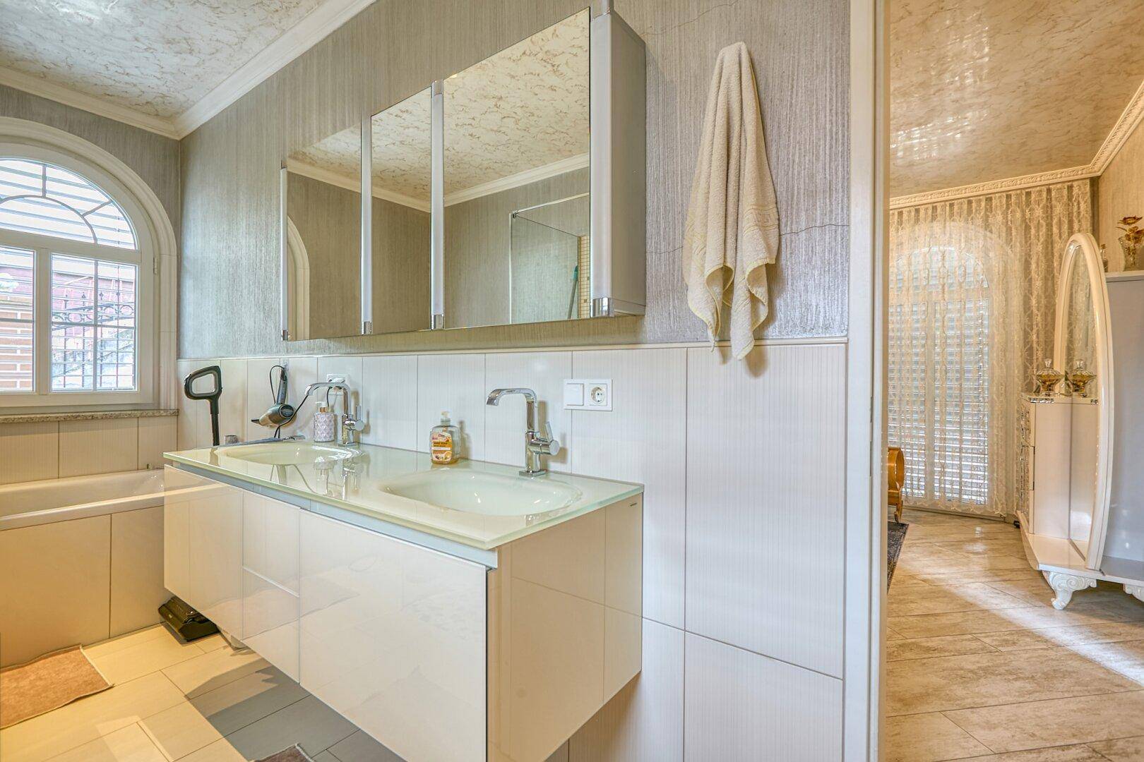 Helles Badezimmer mit Doppelwaschbecken, Badewanne und Spiegelschrank, mit Fenster.