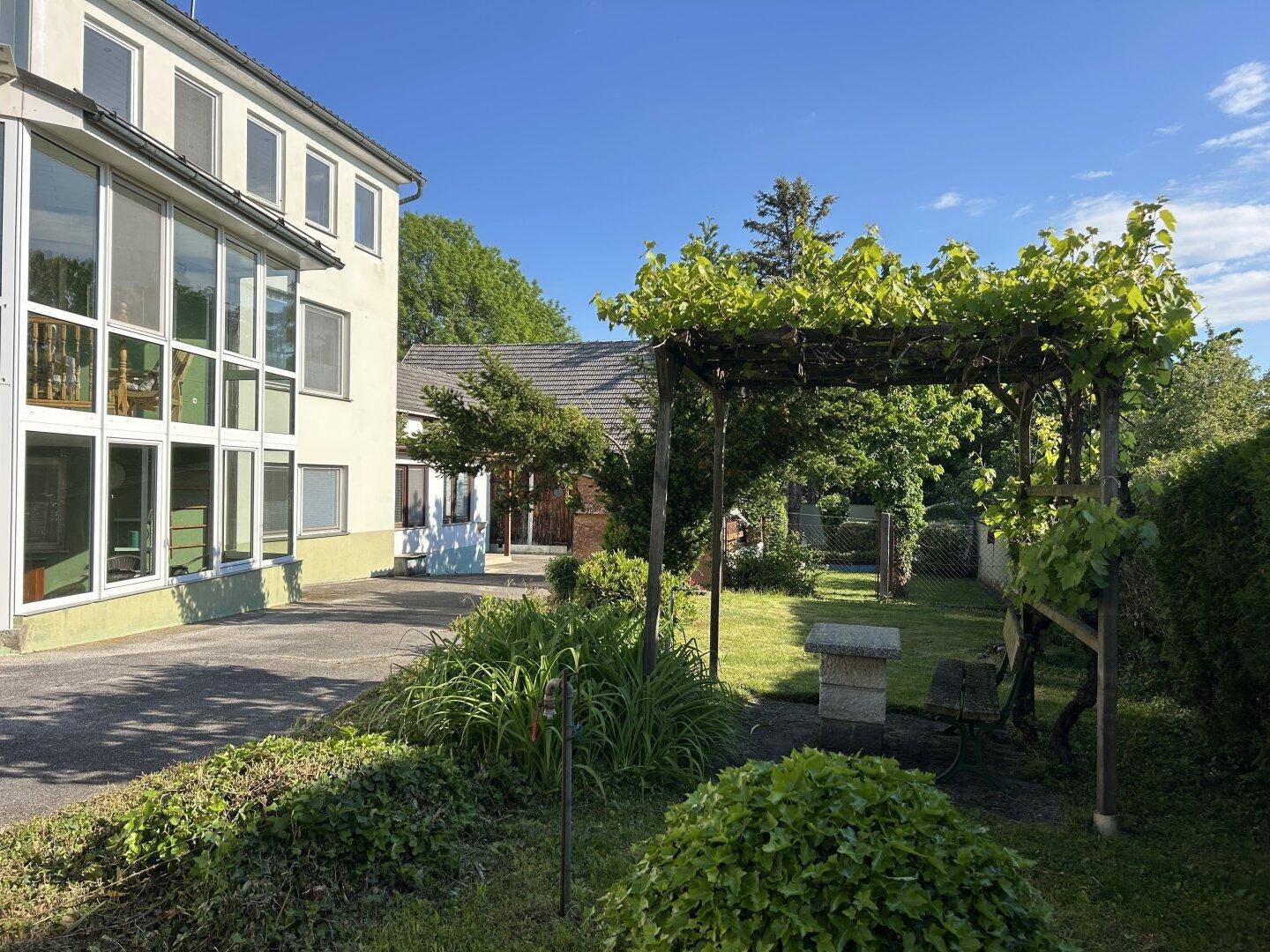 Sonnige Gartenecke mit üppiger Vegetation, Pergola und Blick auf die Gebäude des Anwesens.