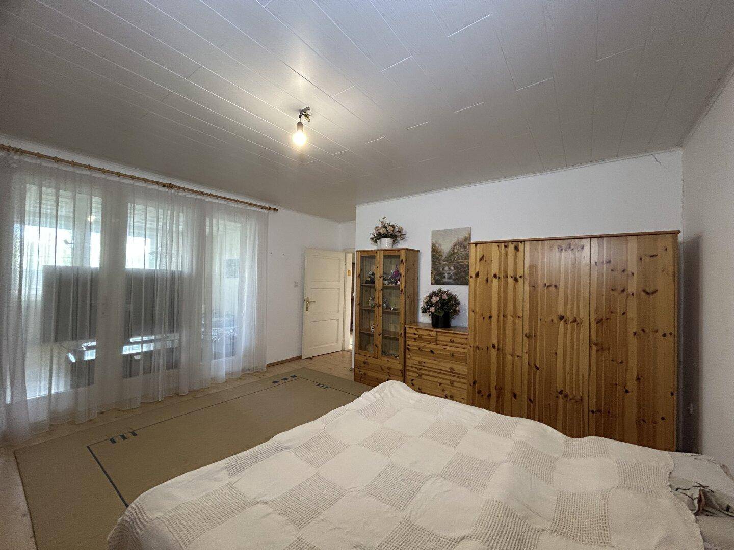 Helles Schlafzimmer mit großem Bett, Holzschränken und Fenstern mit Vorhängen.