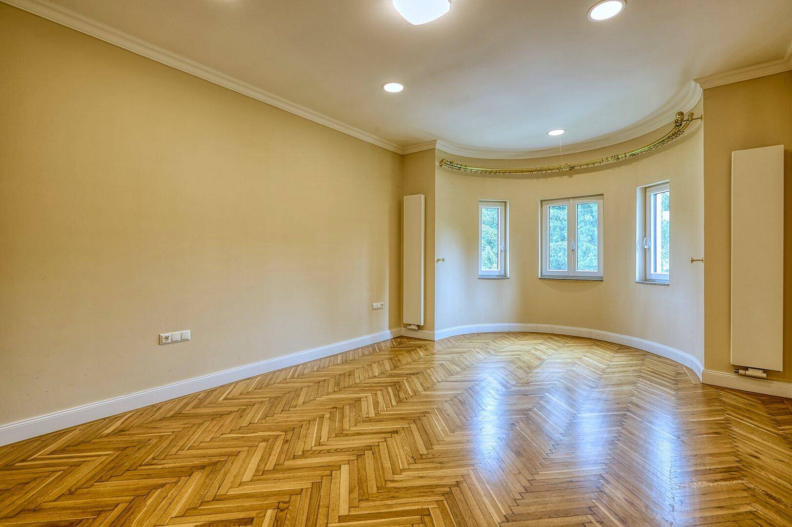 Geräumiges Zimmer mit Fischgrätparkett, beigen Wänden und einem hellen Erkerbereich.