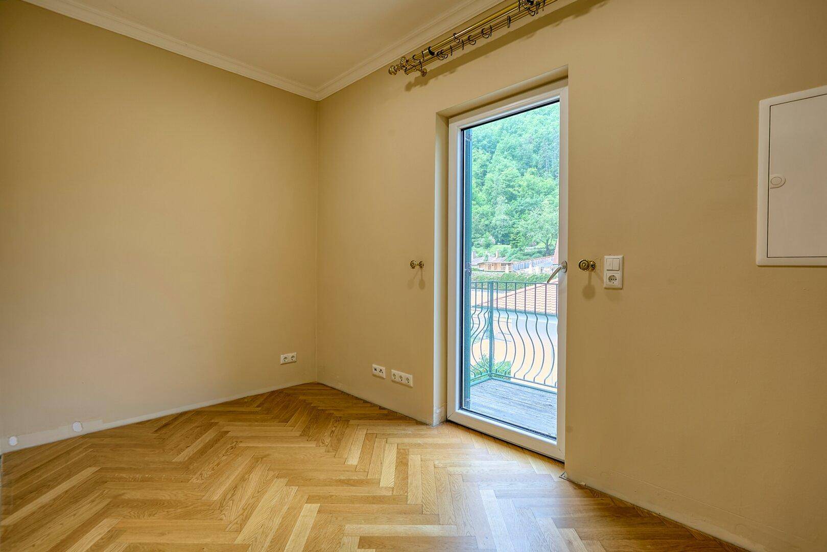 Leeres Zimmer mit Fischgrätparkett und Zugang zu einem kleinen Balkon mit Blick ins Grüne.