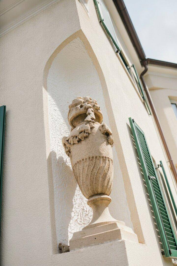 Detailansicht einer Hausfassade mit dekorativer Steinvase in einer Nische und grünen Fensterläden.