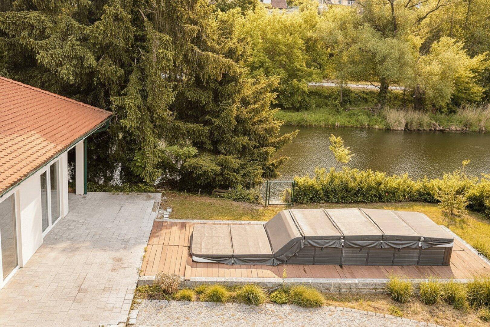 Großer überdachter Whirlpool auf einer Holzterrasse neben einem gepflasterten Bereich und einem See.