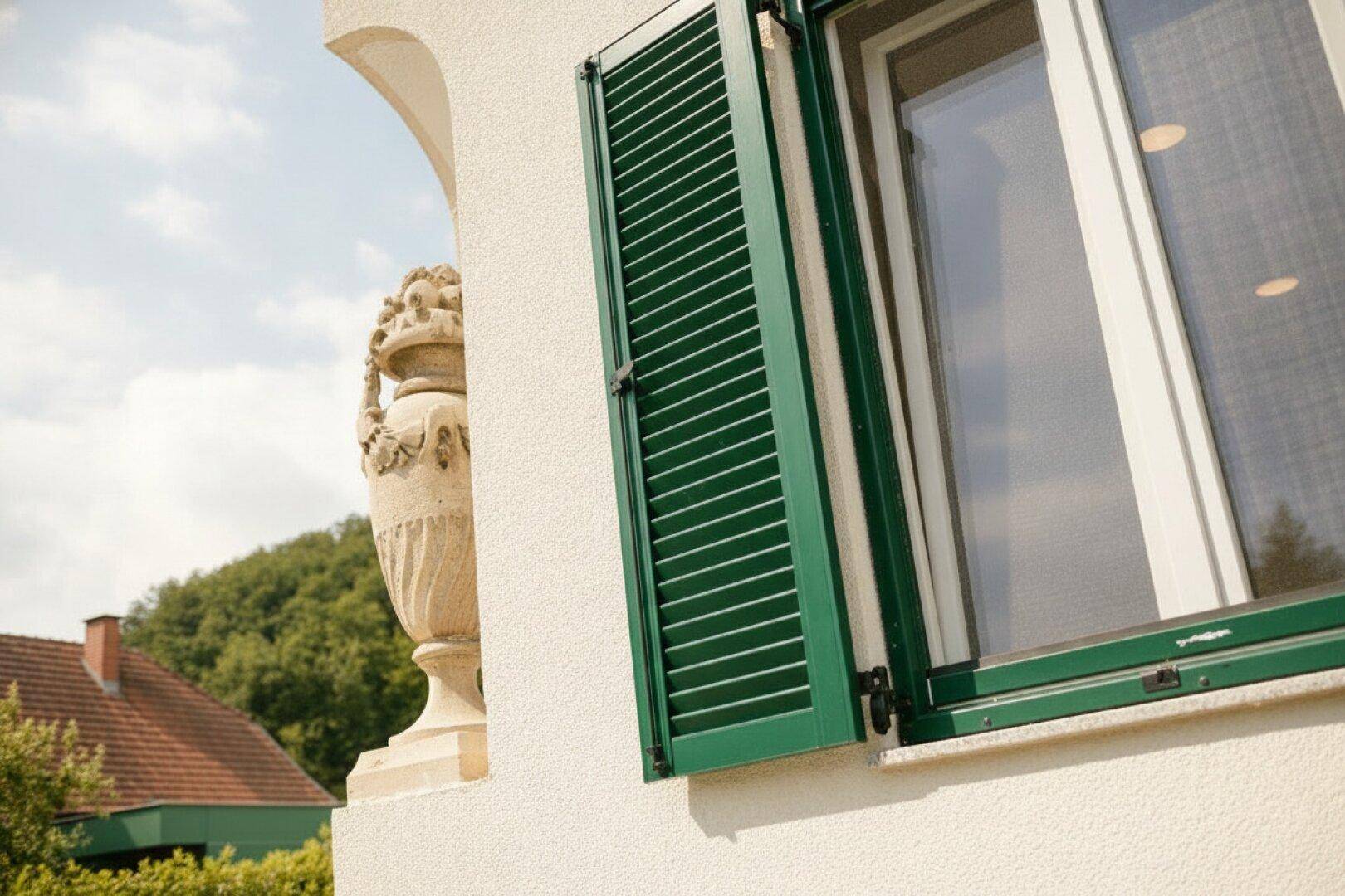Nahaufnahme eines Fensters mit grünen Holzläden und einer dekorativen Steinvase im Hintergrund.
