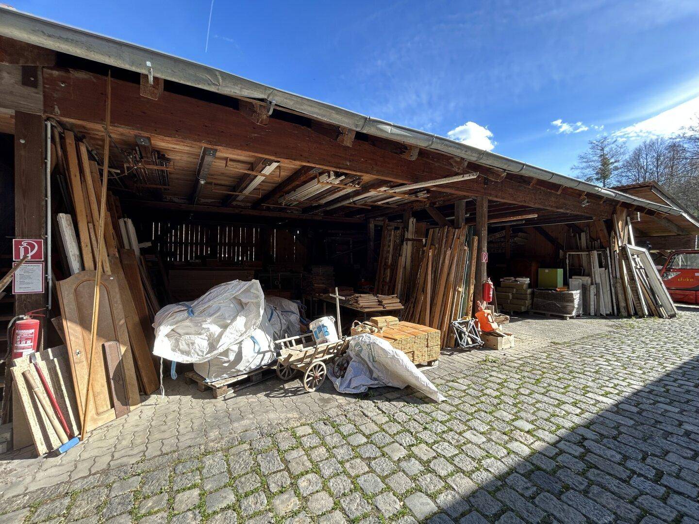 Großer überdachter Lagerbereich mit Holzmaterialien und gepflastertem Boden.