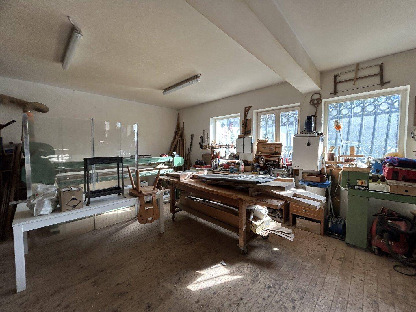 Werkstatt mit Arbeitsflächen, Fenstern und Lagerung für Materialien, gut beleuchtet.