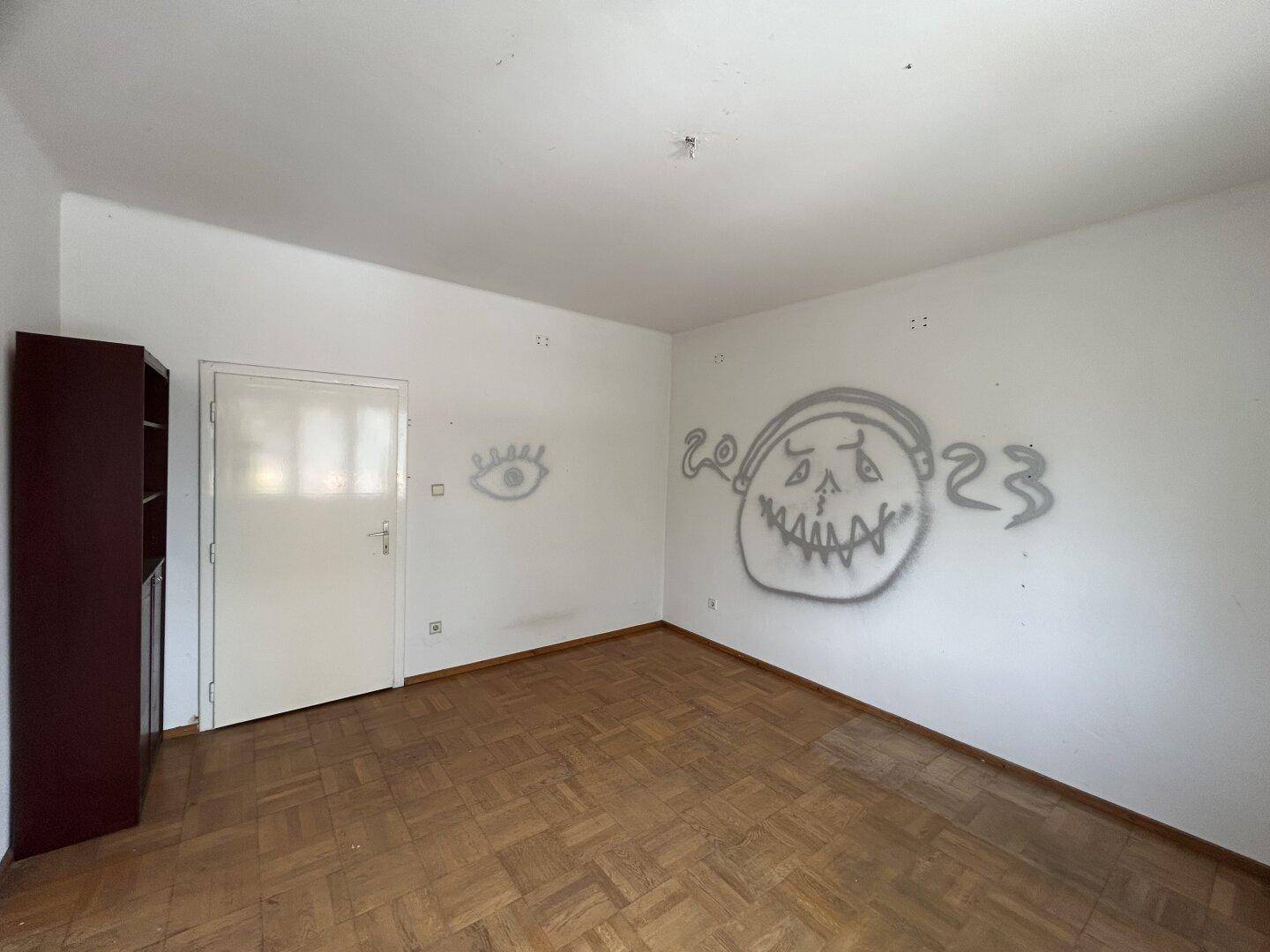 Leeres Zimmer mit Parkettboden, weißen Wänden und Graffiti, bereit zur Renovierung.