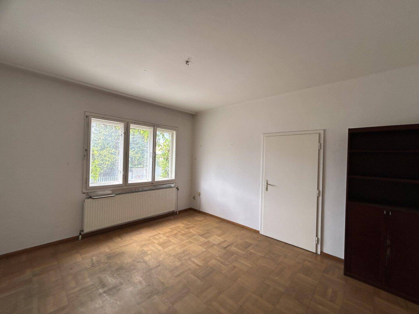 Leeres Zimmer mit Parkettboden, Fenster und Heizkörper, bietet Blick ins Grüne.