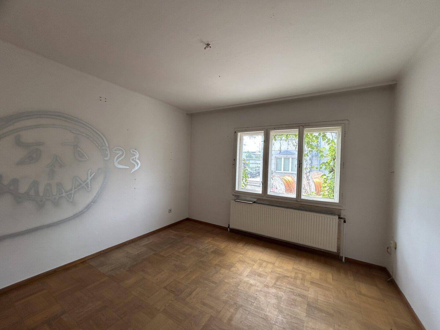 Leeres Zimmer mit Parkettboden, Graffiti an der Wand und Fenster mit Blick nach draußen.