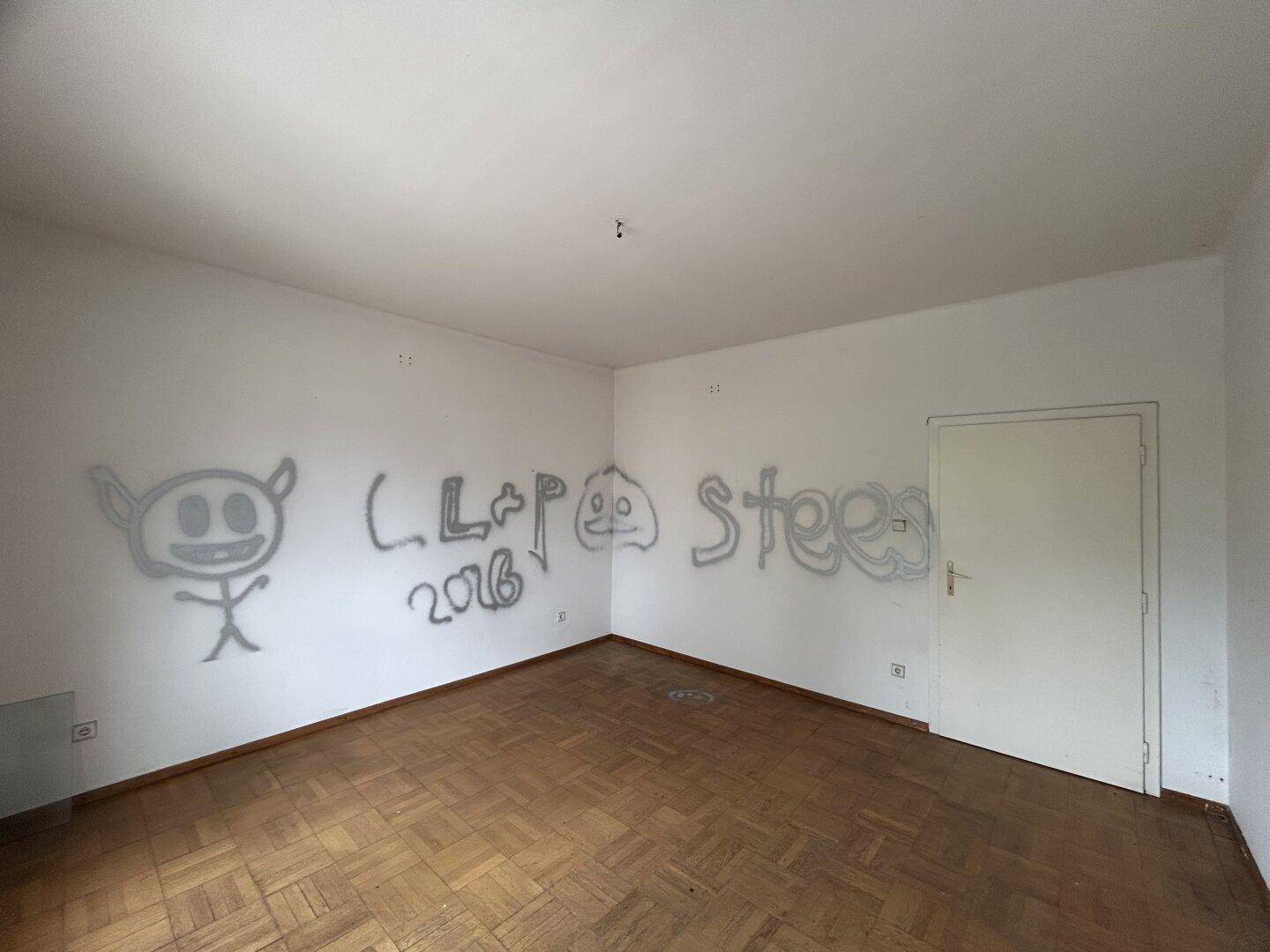 Leeres Zimmer mit Parkettboden und Graffiti an der Wand, bereit zur Renovierung.