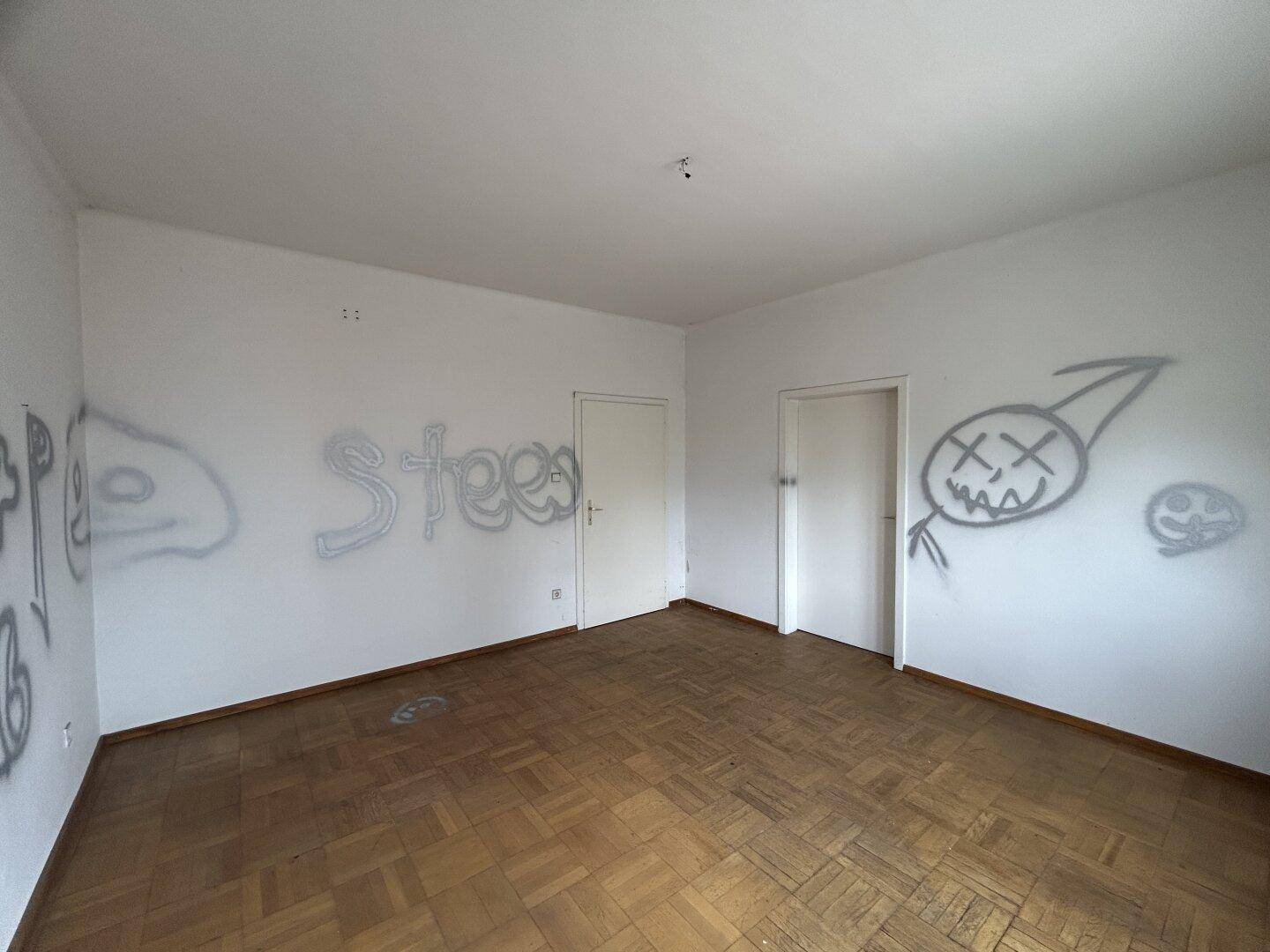 Leeres Zimmer mit Parkettboden, Graffiti an den Wänden und zwei Türen.