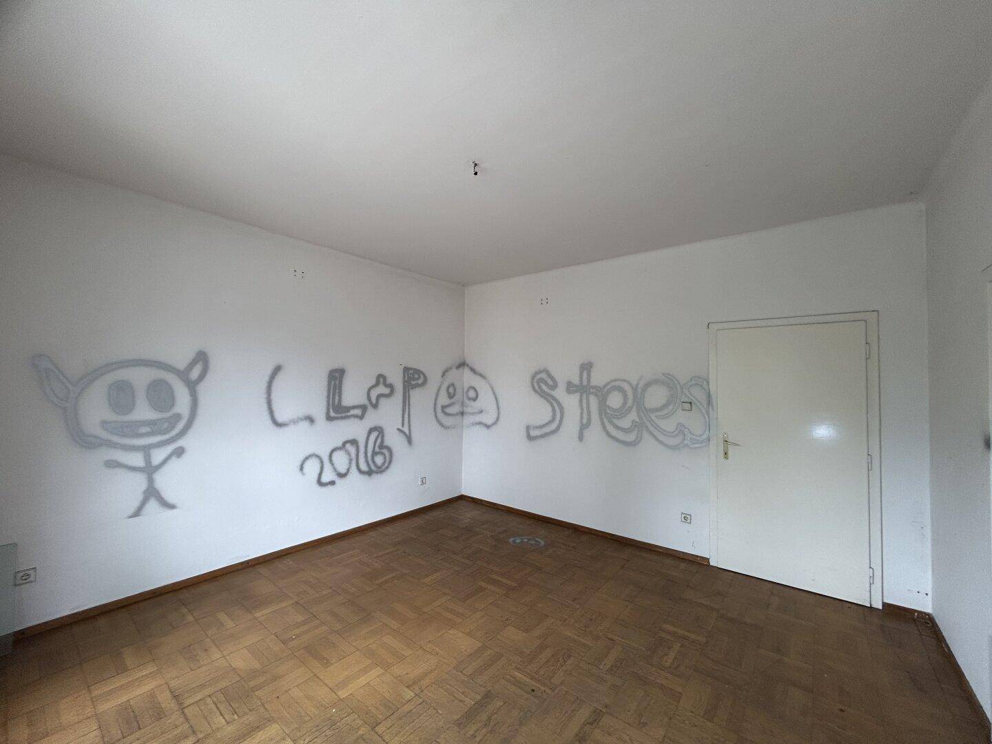 Leeres Zimmer mit Parkettboden und Graffiti an der Wand, bereit zur Renovierung.
