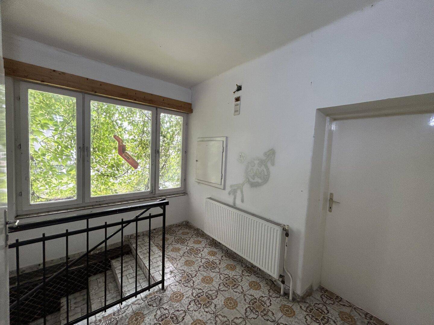 Flur mit gemusterten Fliesen, Treppe und Fenster mit Blick auf grüne Bäume.