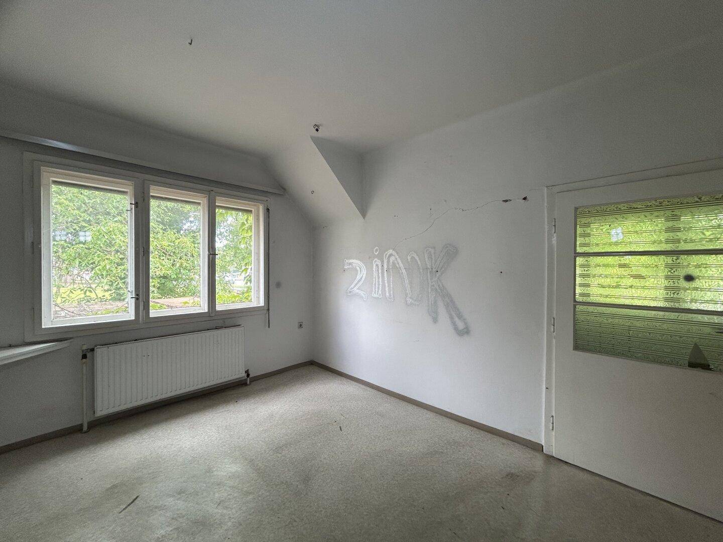 Leeres Zimmer mit Teppichboden, Fenster und schräger Decke, mit Graffiti an der Wand.