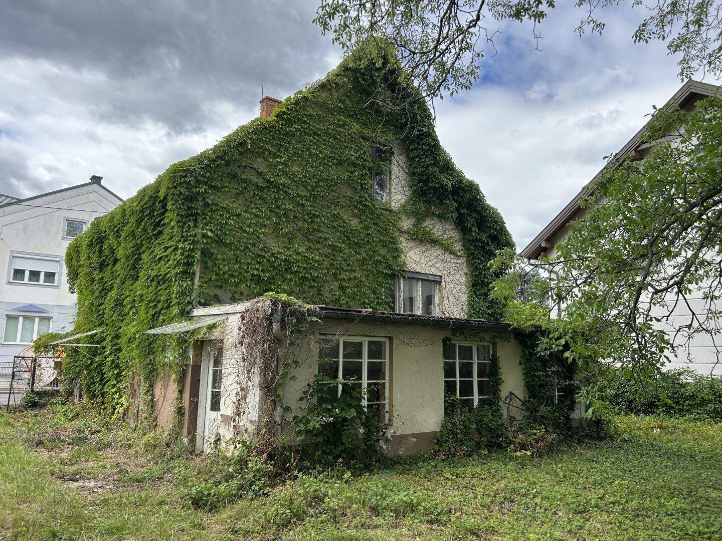 Seitenansicht eines stark bewachsenen Hauses mit verwildertem Garten und angrenzendem Gebäude.