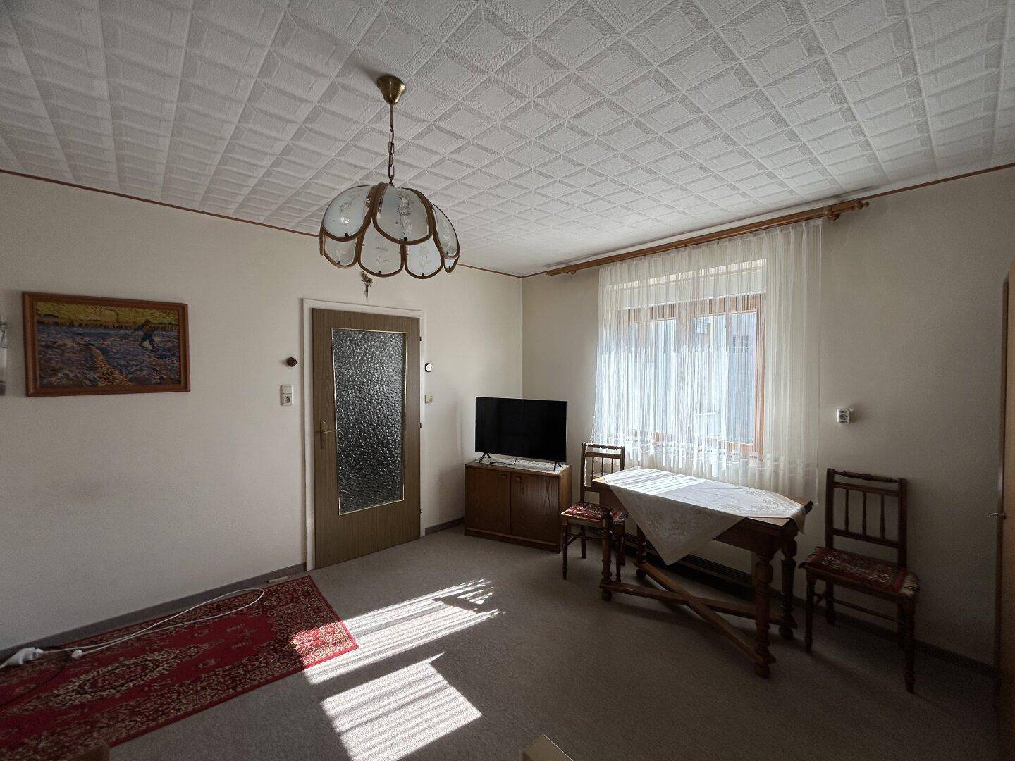 Wohnzimmer mit Tisch, Fernseher und Fenster, bietet viel Tageslicht und eine gemütliche Atmosphäre.