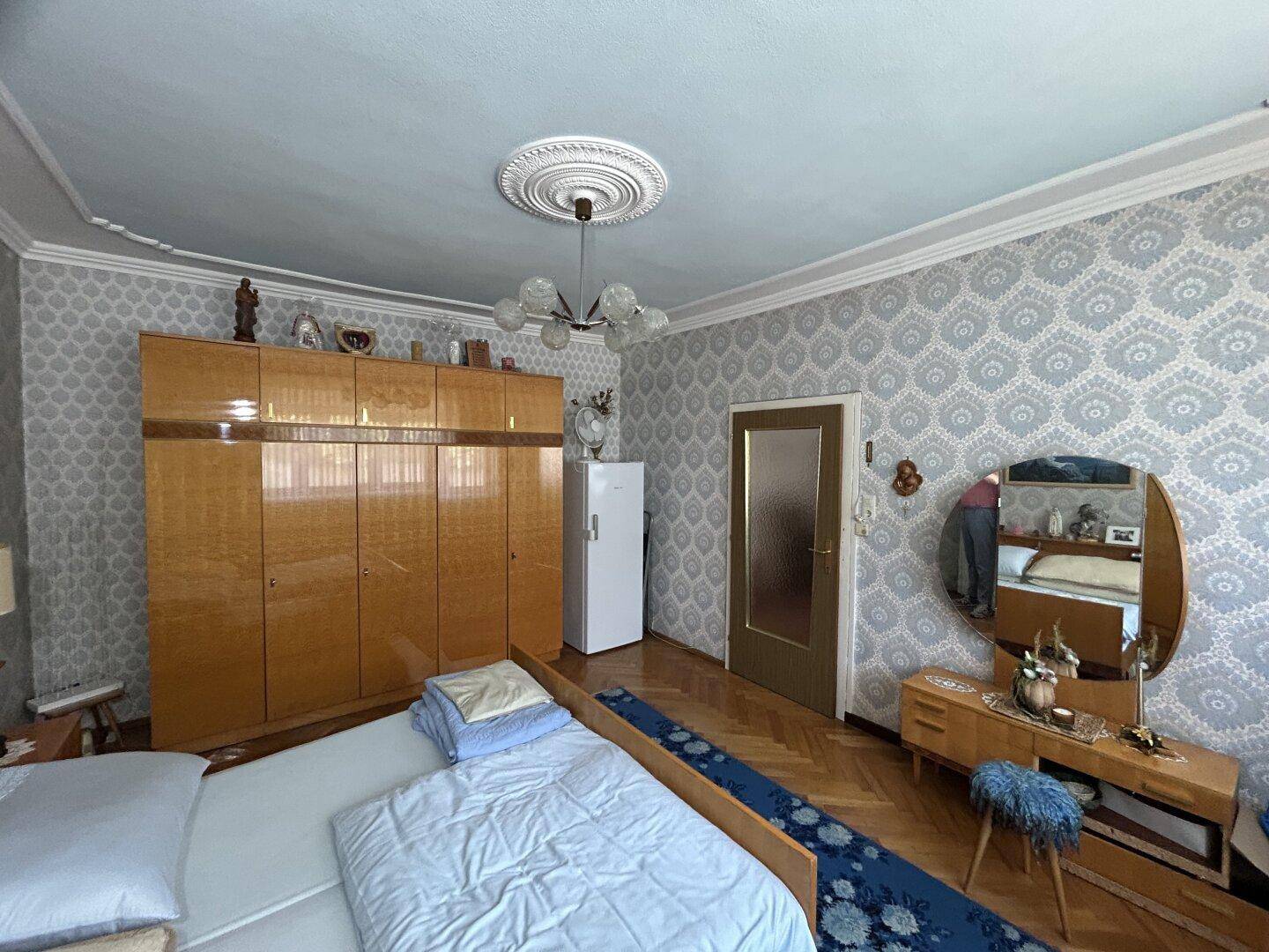 Schlafzimmer im Vintage-Stil mit gemusterter Tapete, Holzschrank und Parkettboden.