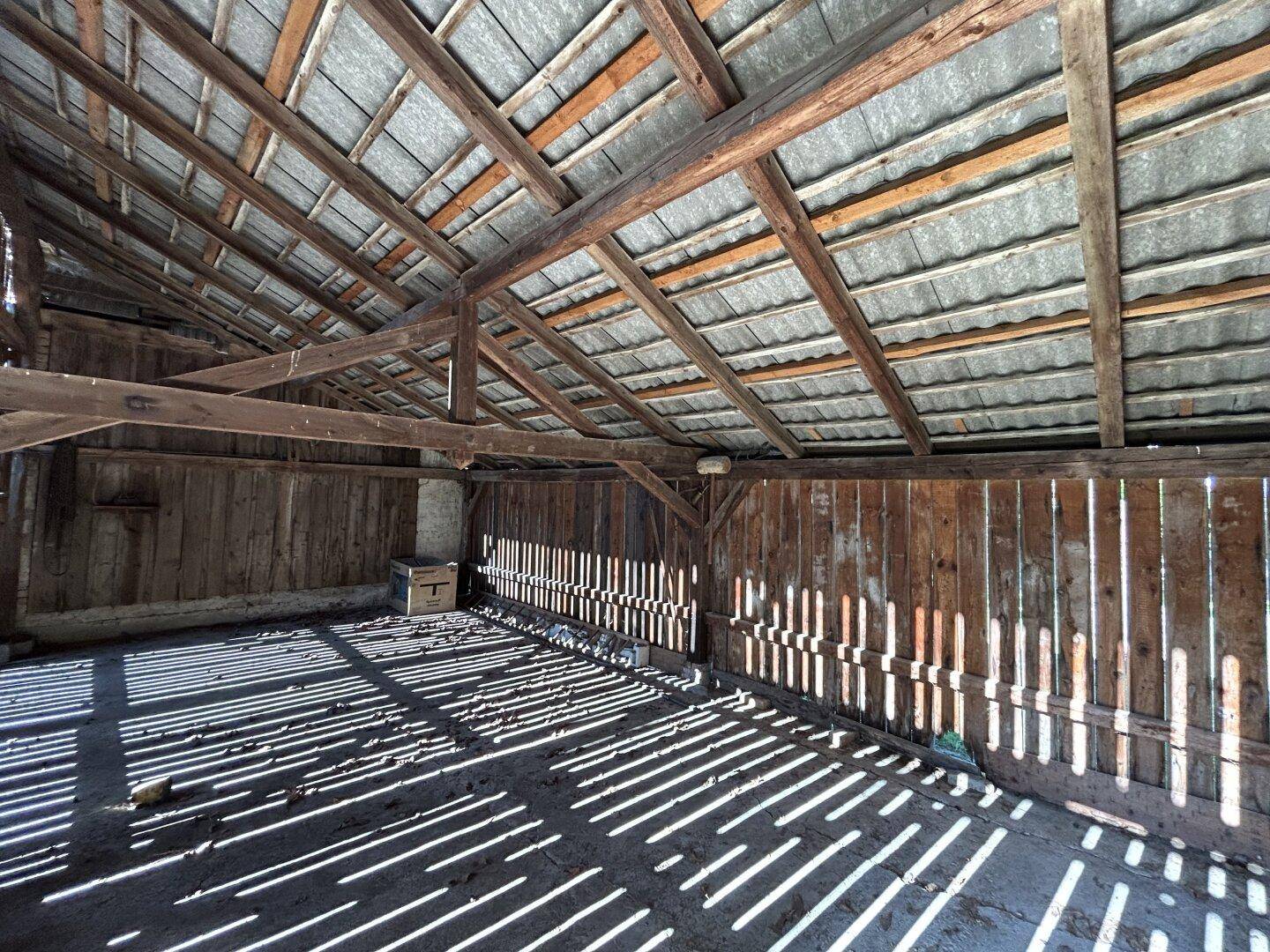 Innenansicht einer Scheune mit Holzwänden, Dachkonstruktion und Streifen von Sonnenlicht.