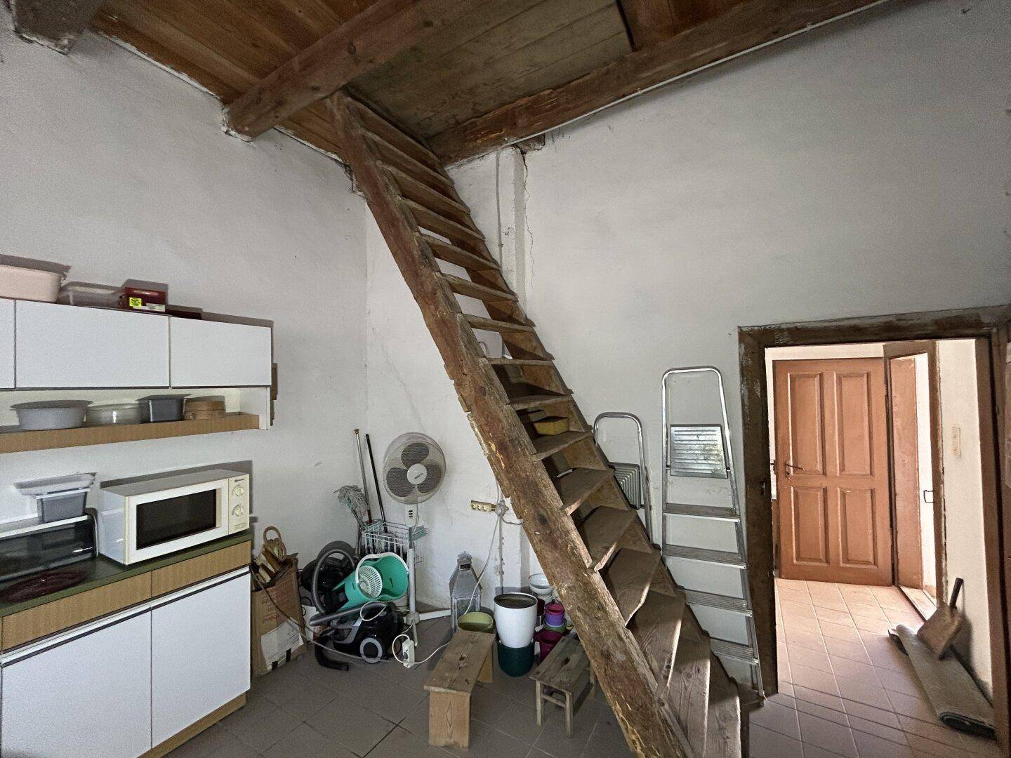 Einfache Küche mit Holztreppe zum Obergeschoss und Blick auf den Eingangsbereich.