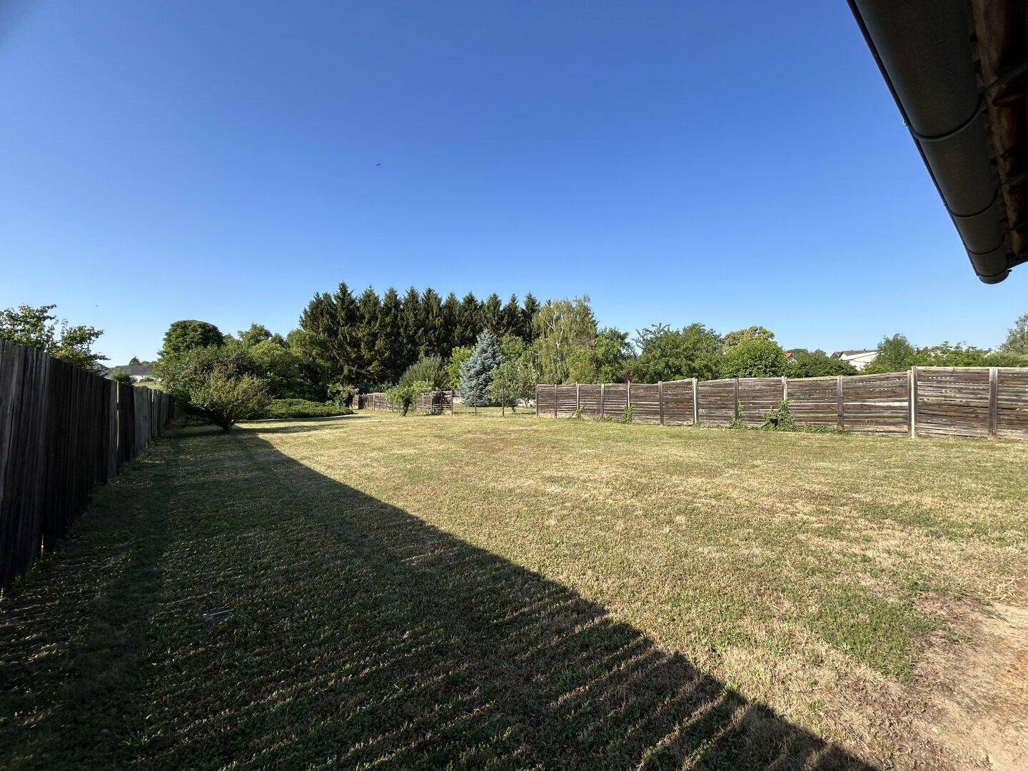 Weitläufiger Garten mit Rasenfläche, umzäunt von einem Holzzaun und Bäumen im Hintergrund.
