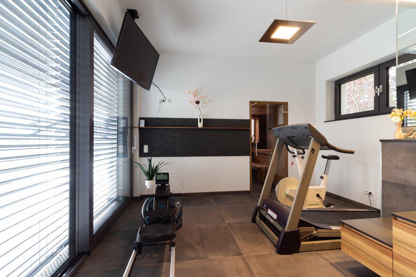 Moderner Fitnessraum mit Trainingsgeräten, Fernseher und Zugang zu einer Sauna.