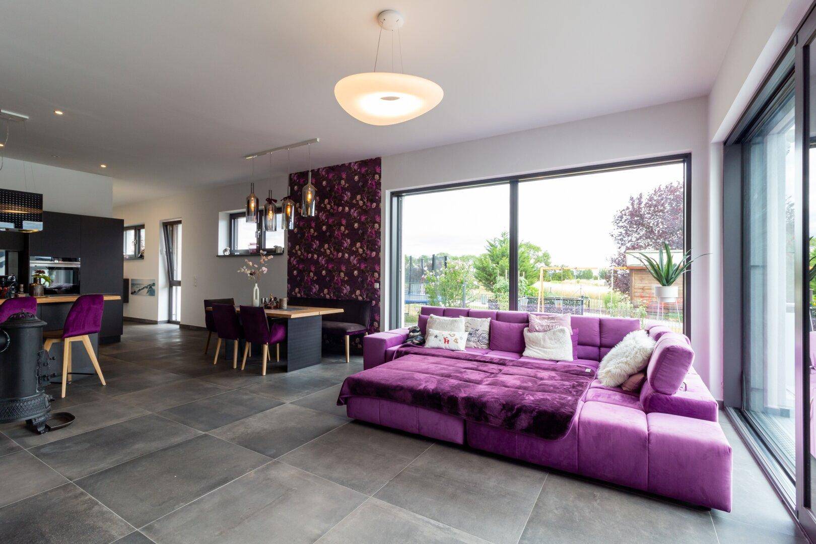 Helles Wohnzimmer mit großem lila Sofa, dunklen Fliesen und bodentiefen Fenstern.