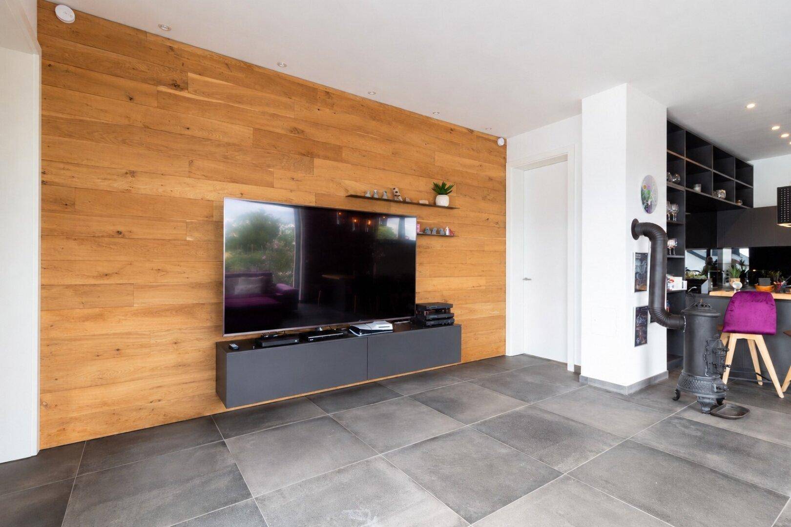 Wohnbereich mit Holzwand, großem Fernseher und dunklen Fliesen, offen zur Küche.