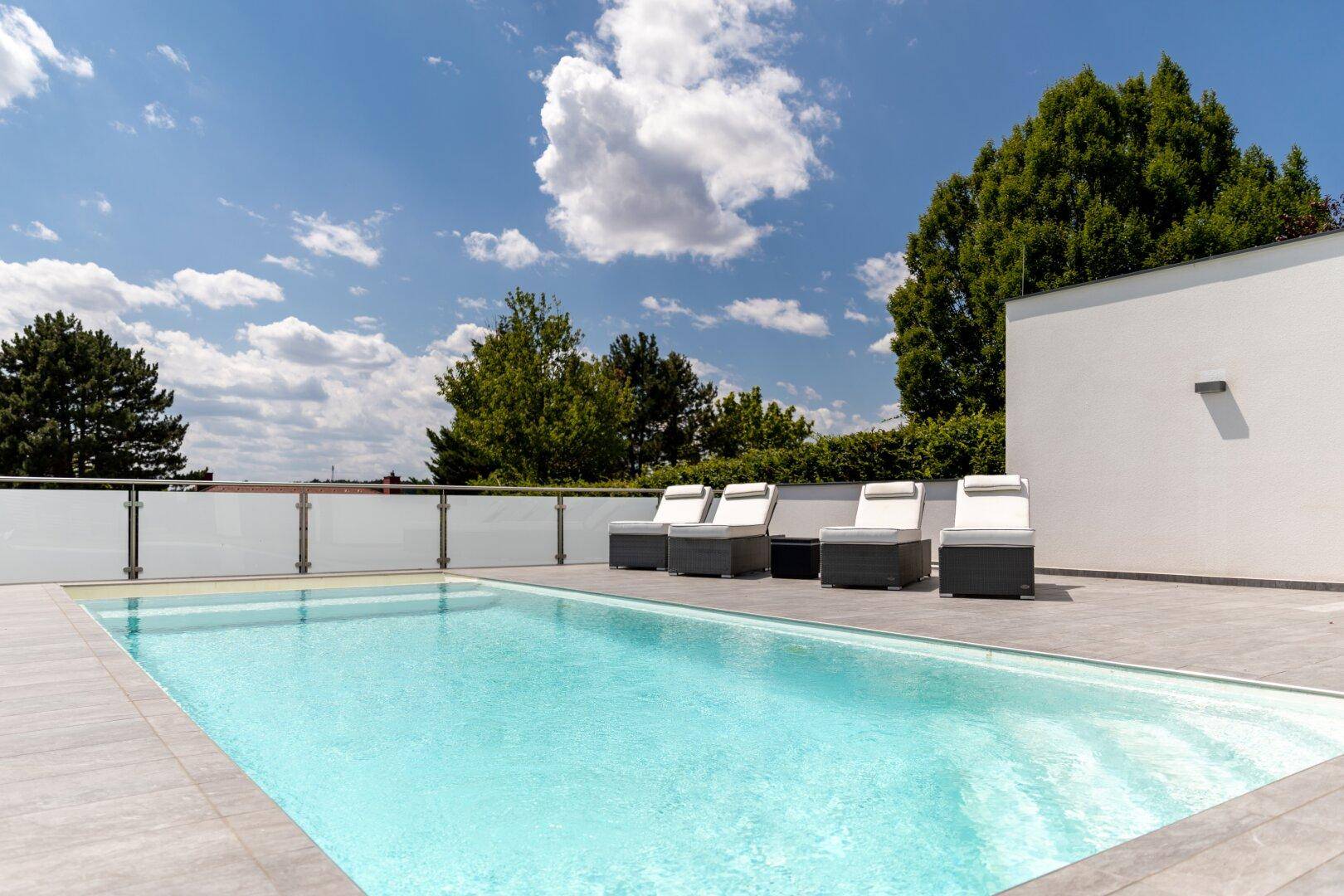 Weitläufiger Poolbereich mit mehreren Sonnenliegen unter blauem Himmel und grüner Umgebung.