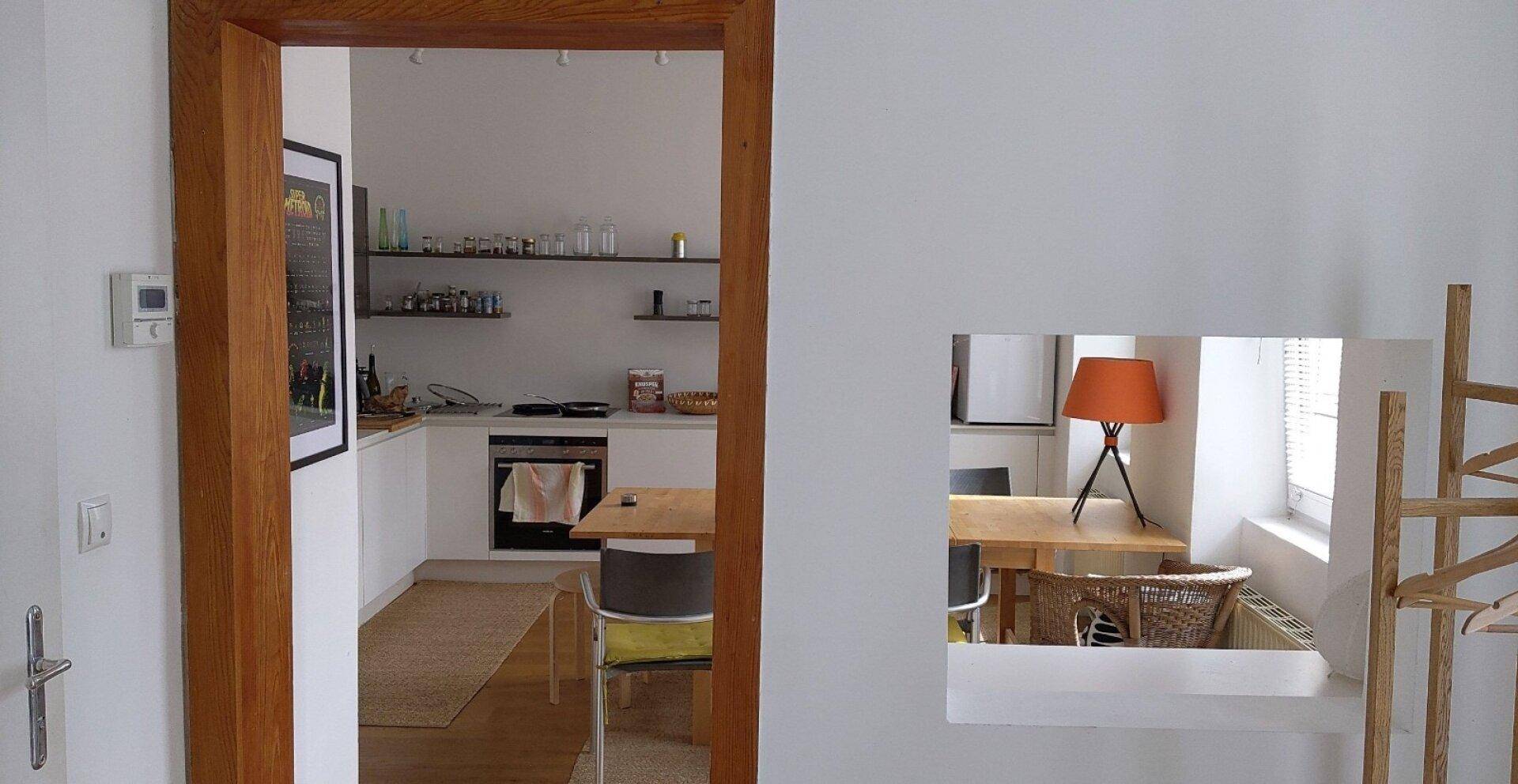 Blick durch eine Holztür in eine moderne Küche mit Einbaugeräten und Essbereich.
