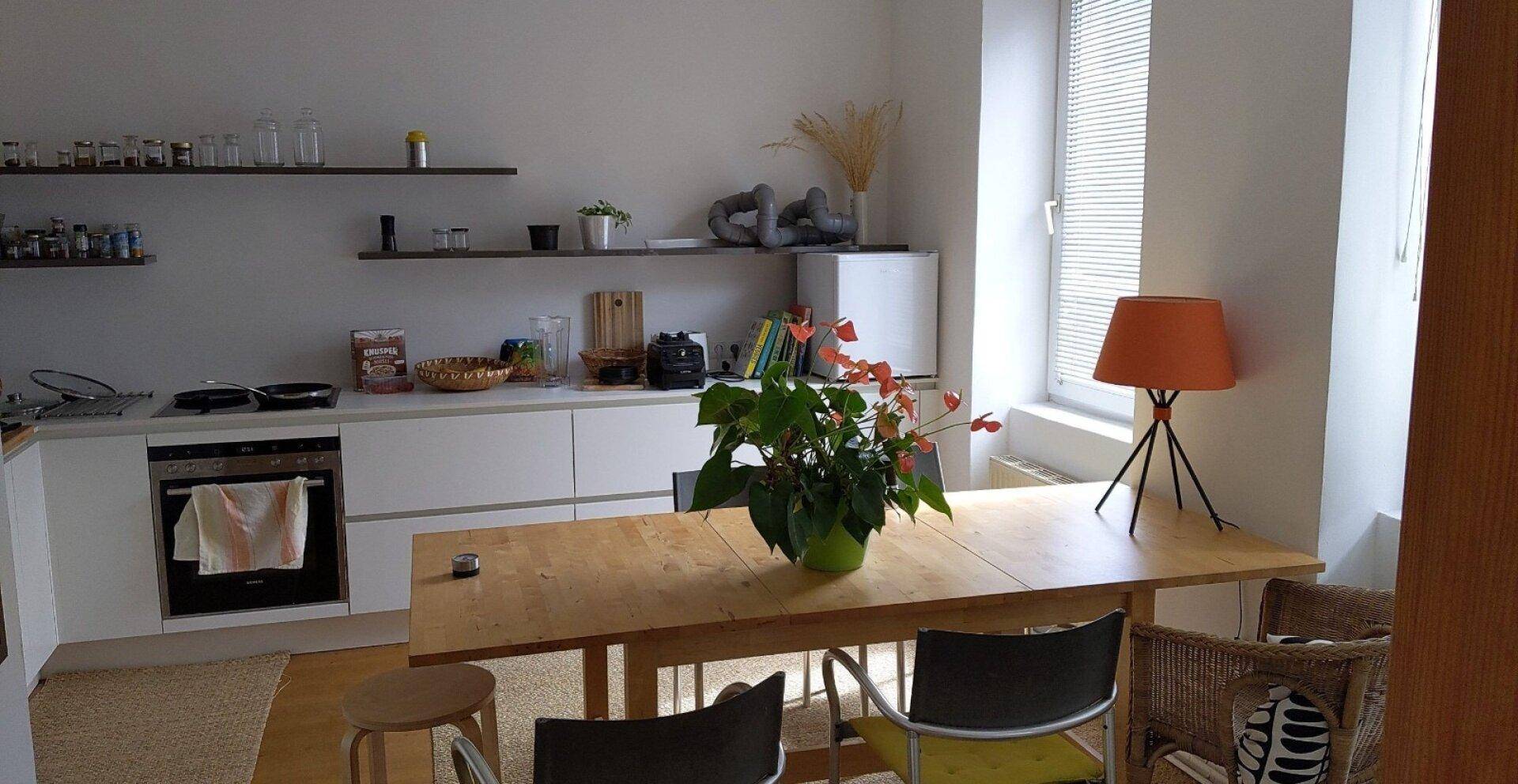 Helle Küche mit weißer Einbauküche, Esstisch und Fenster mit Jalousien.