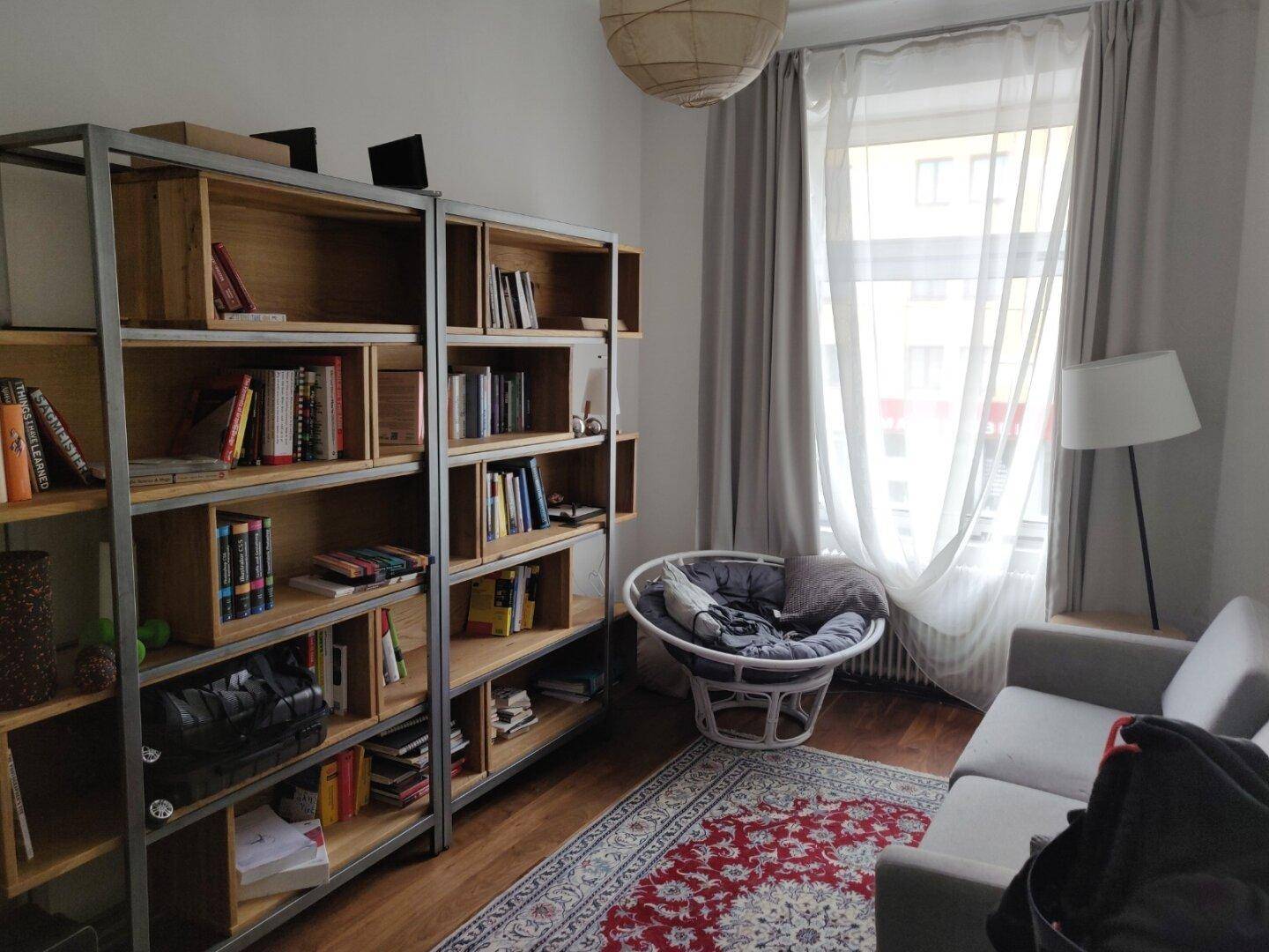Wohnzimmer mit Bücherregalen, Sofa, Sessel und einem Fenster mit Vorhängen.